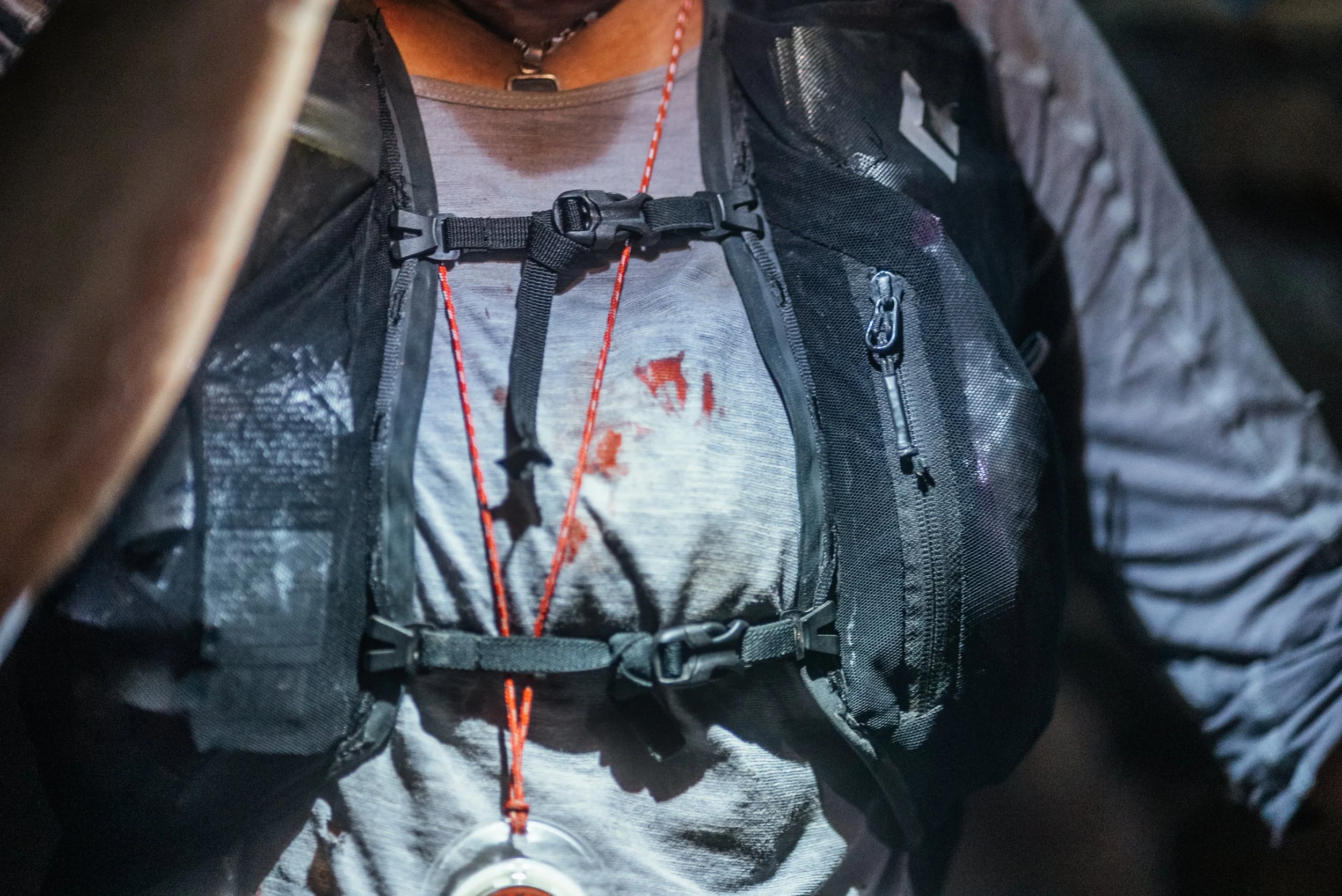 Close-up of a person wearing a black backpack with multiple zippers and straps, over a gray t-shirt, and a red lanyard with a round badge or medallion hanging from it.