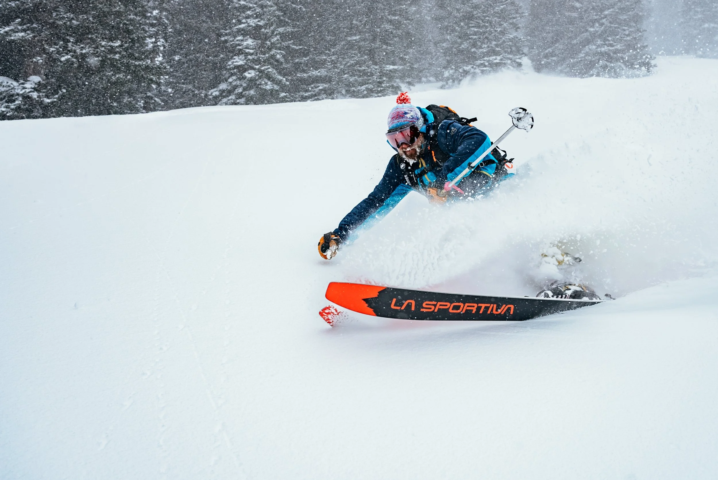 A man wearing winter gear and goggles is skiing down a snowy slope, kicking up snow as he turns.