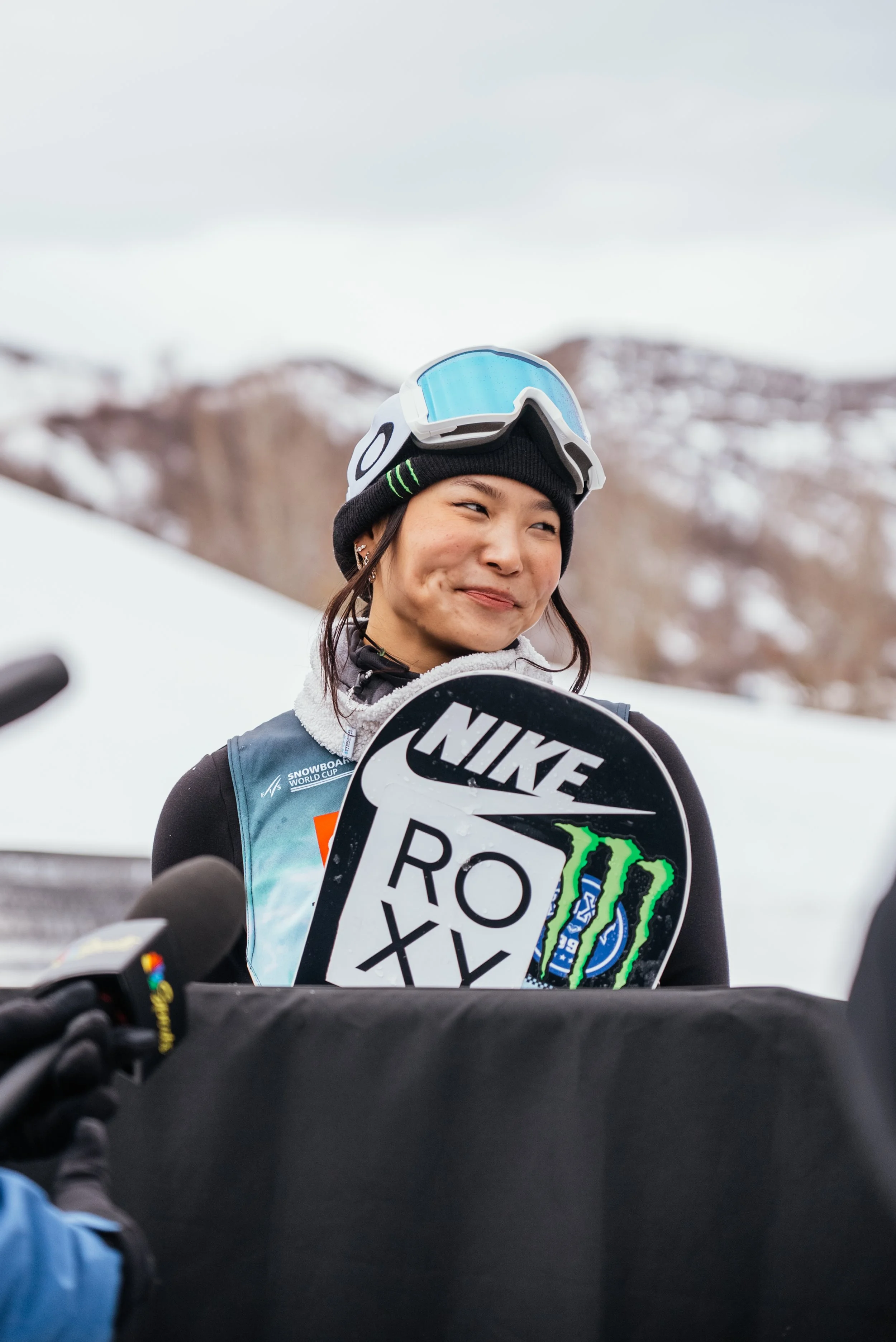 A female snowboarder holding a snowboard with Nike, Monster Energy, and other logos during a snow competition, outdoors with snow-covered mountains in the background.