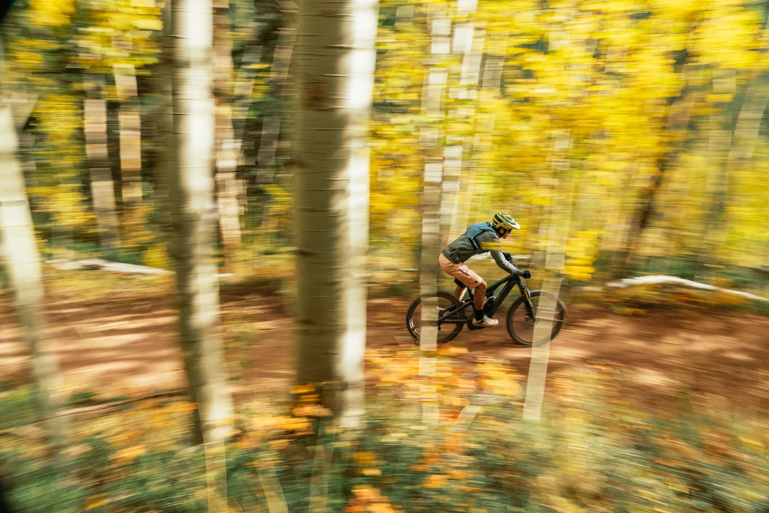 A person mountain biking through an autumn forest with yellow leaves, wearing a helmet, black jacket, and pink shorts.