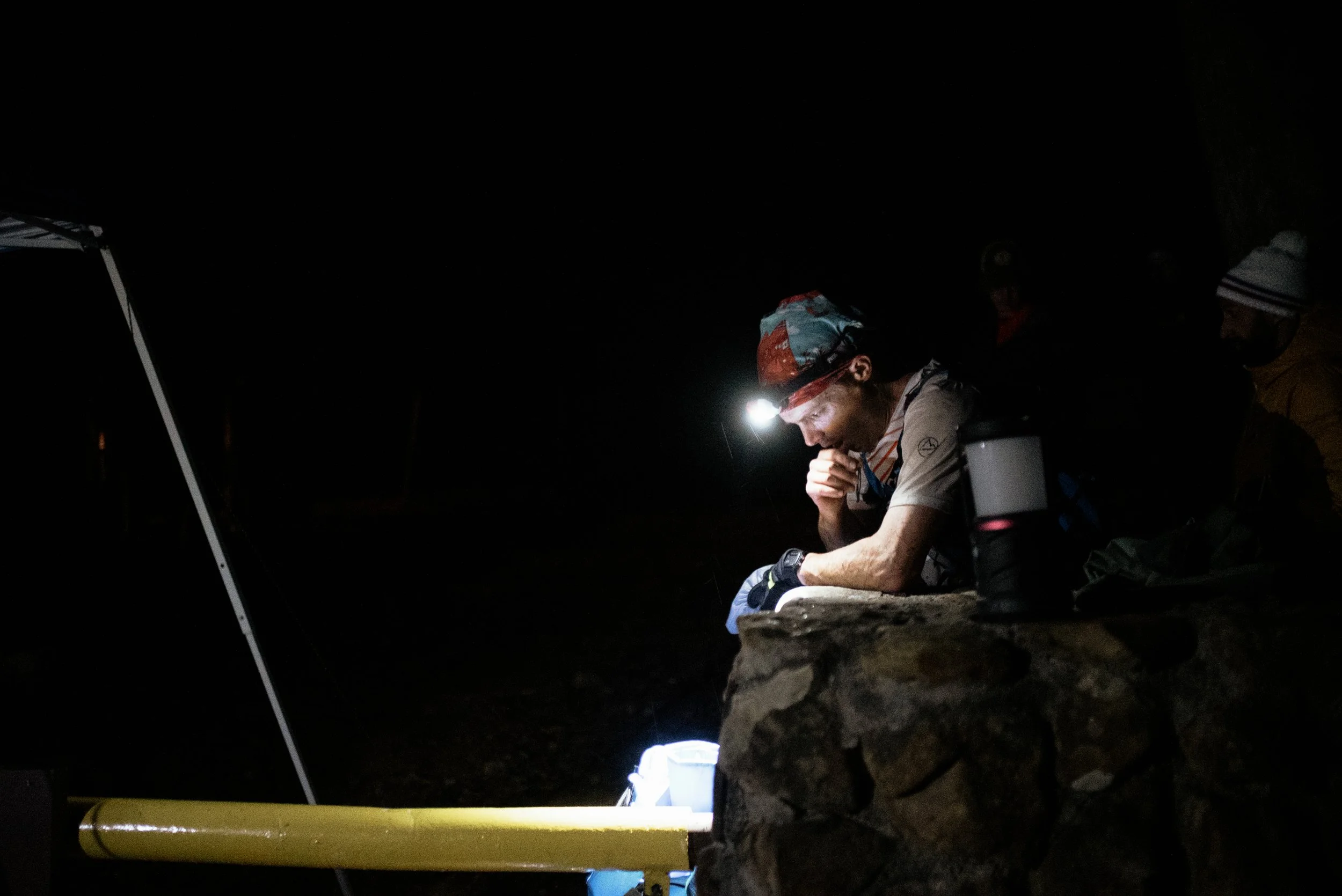 Person sitting on stone wall at night using headlamp, surrounded by dark environment with camping gear.