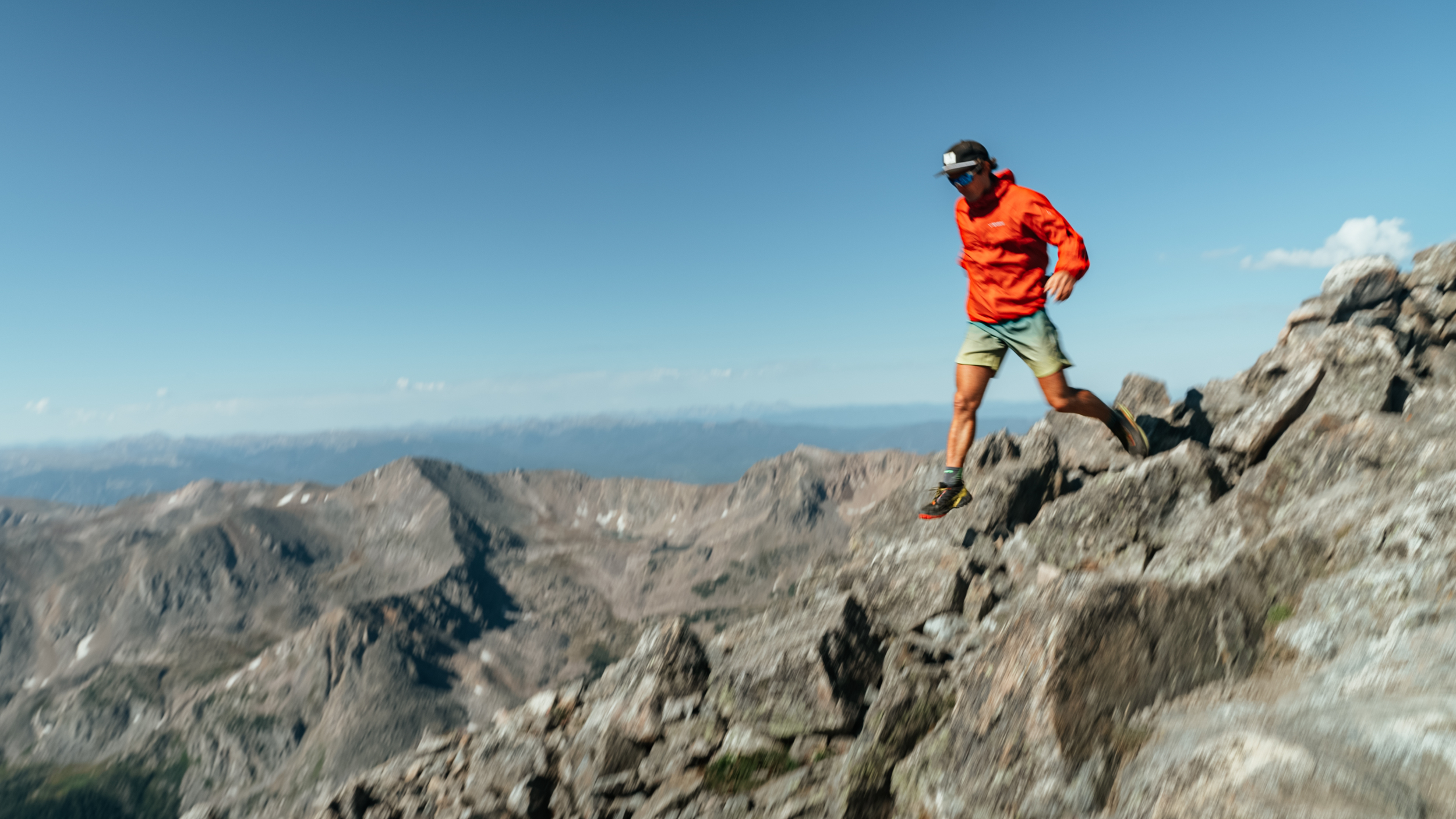 A person wearing an orange jacket, green shorts, and a black cap with sunglasses is hiking down rocky mountain terrain under a blue sky.