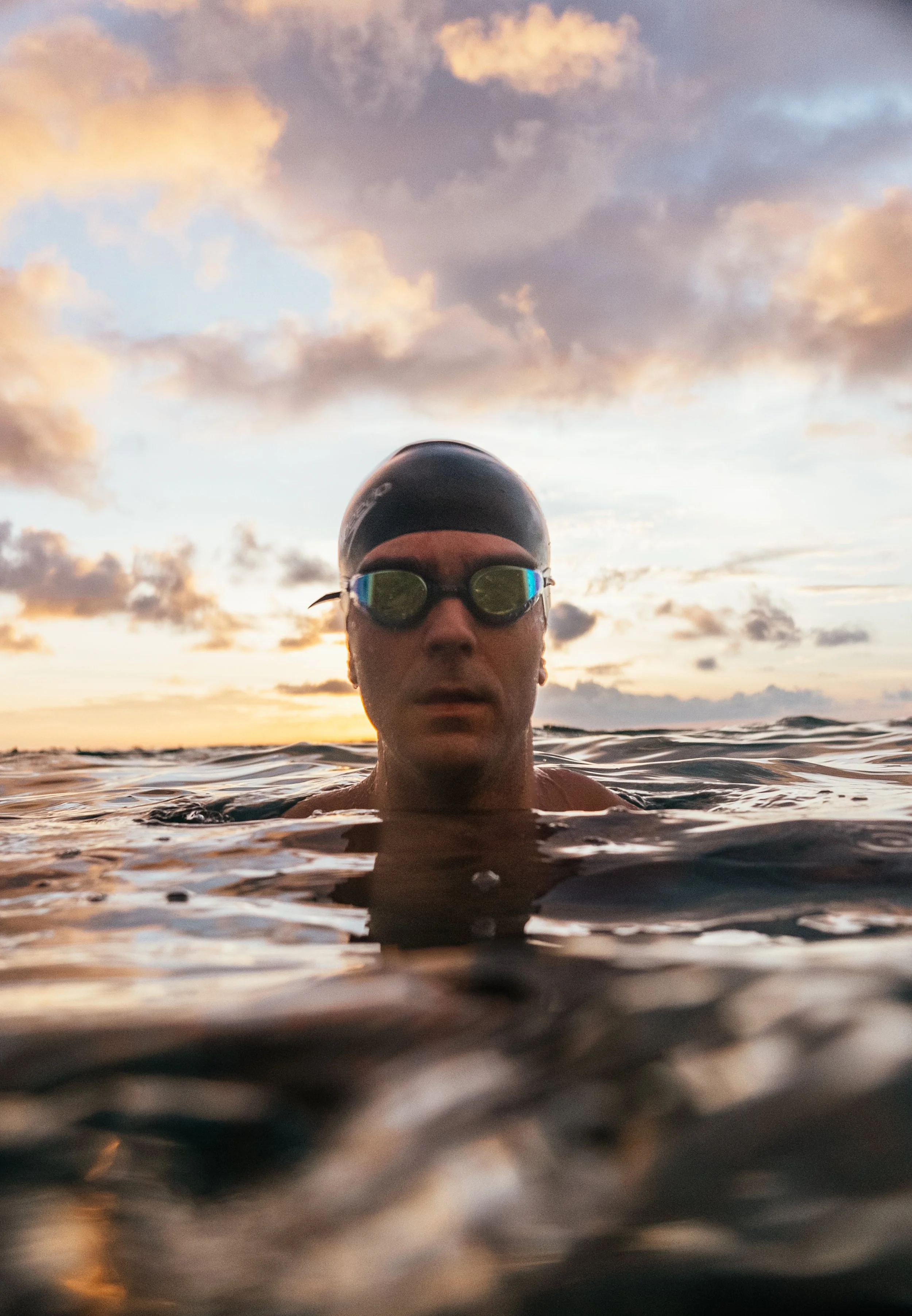 A swimmer wearing goggles and a swim cap in open water at sunrise or sunset, with a partly cloudy sky in the background.