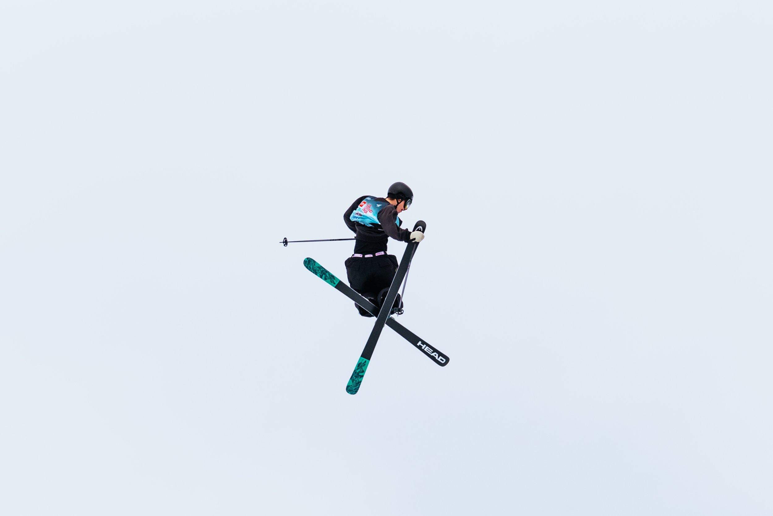 Skier mid-air performing a stunt against a clear sky, wearing a helmet and winter gear, skis and poles visible.