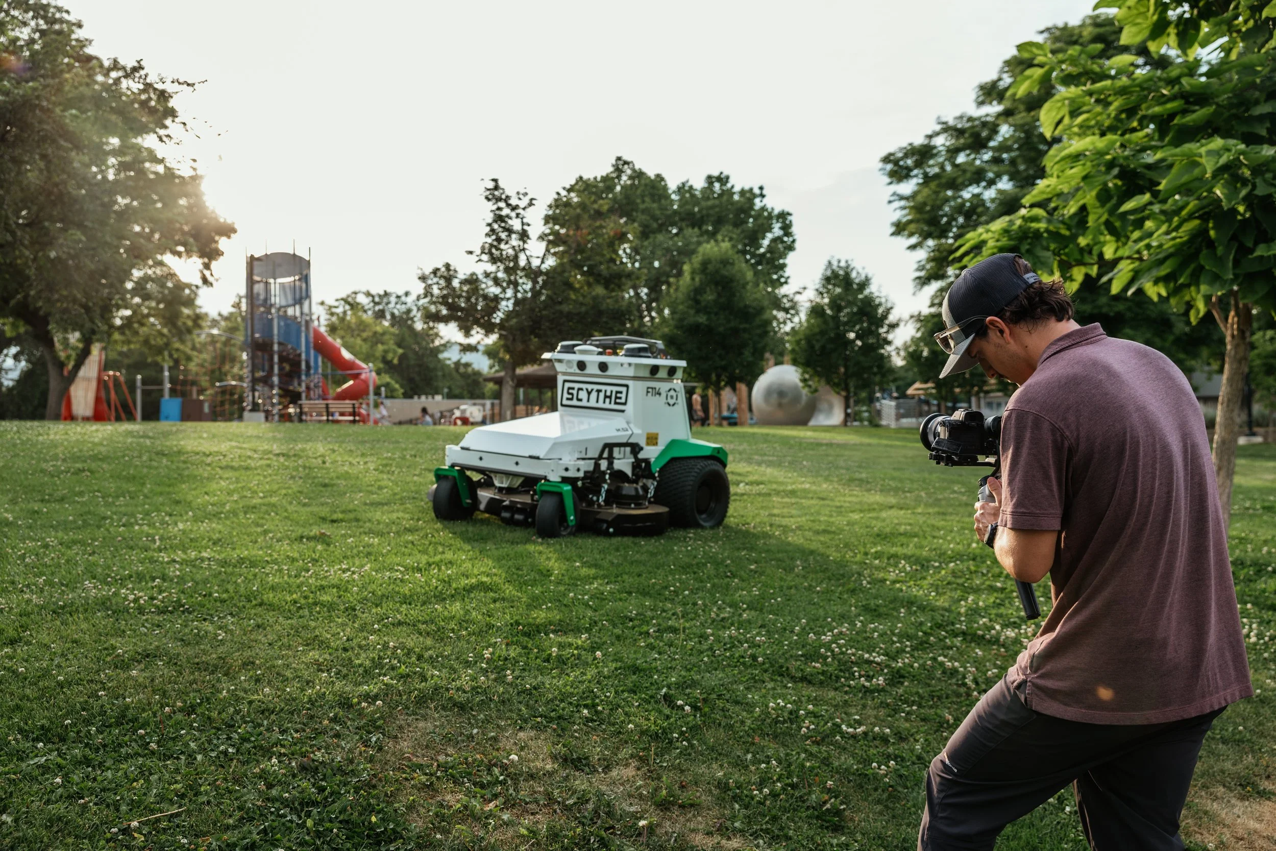 A man with a camera films an autonomous lawnmower on a grassy park with playground equipment and trees in the background.