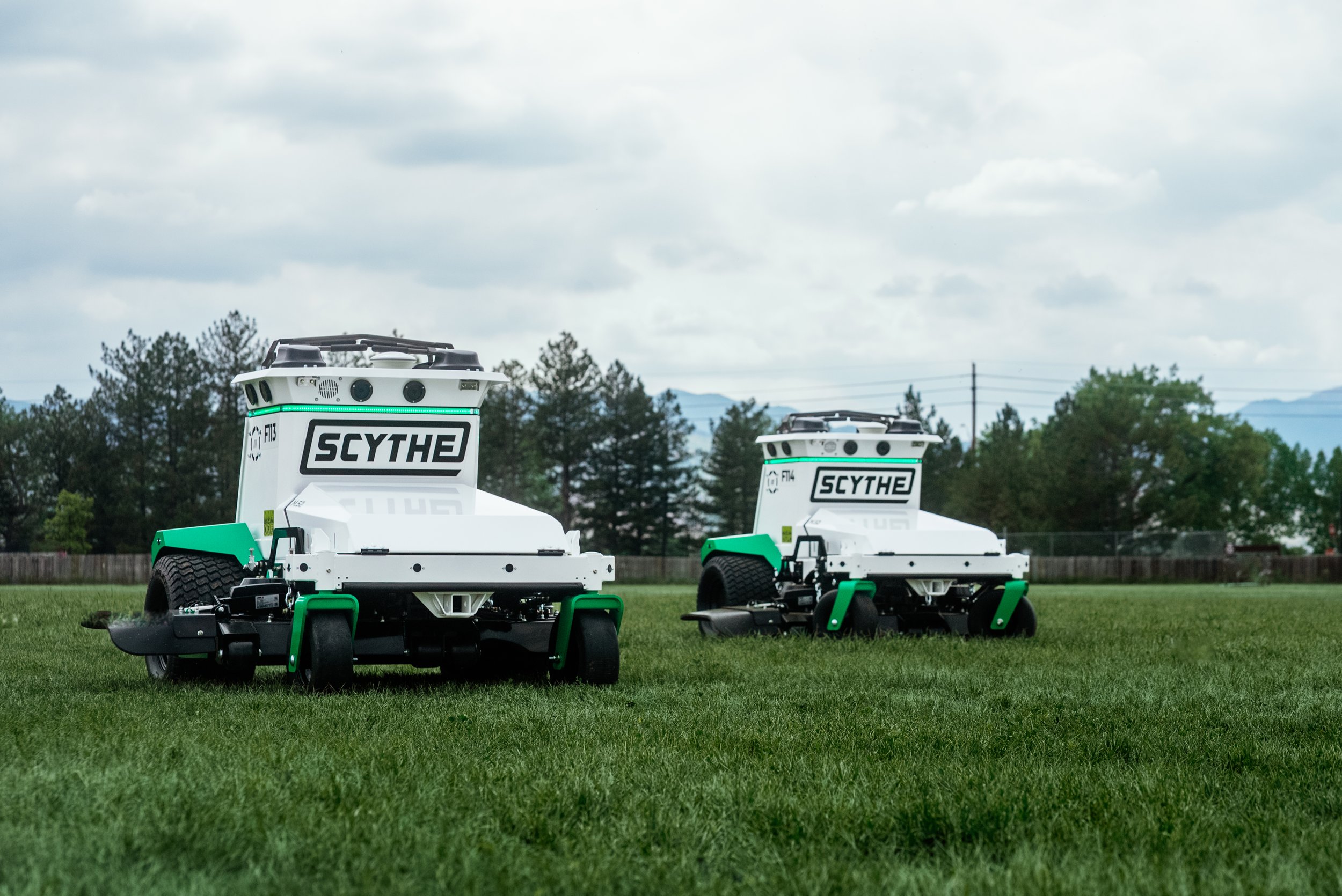 Two white and green autonomous lawn-mowing robots on a grassy field under a cloudy sky.