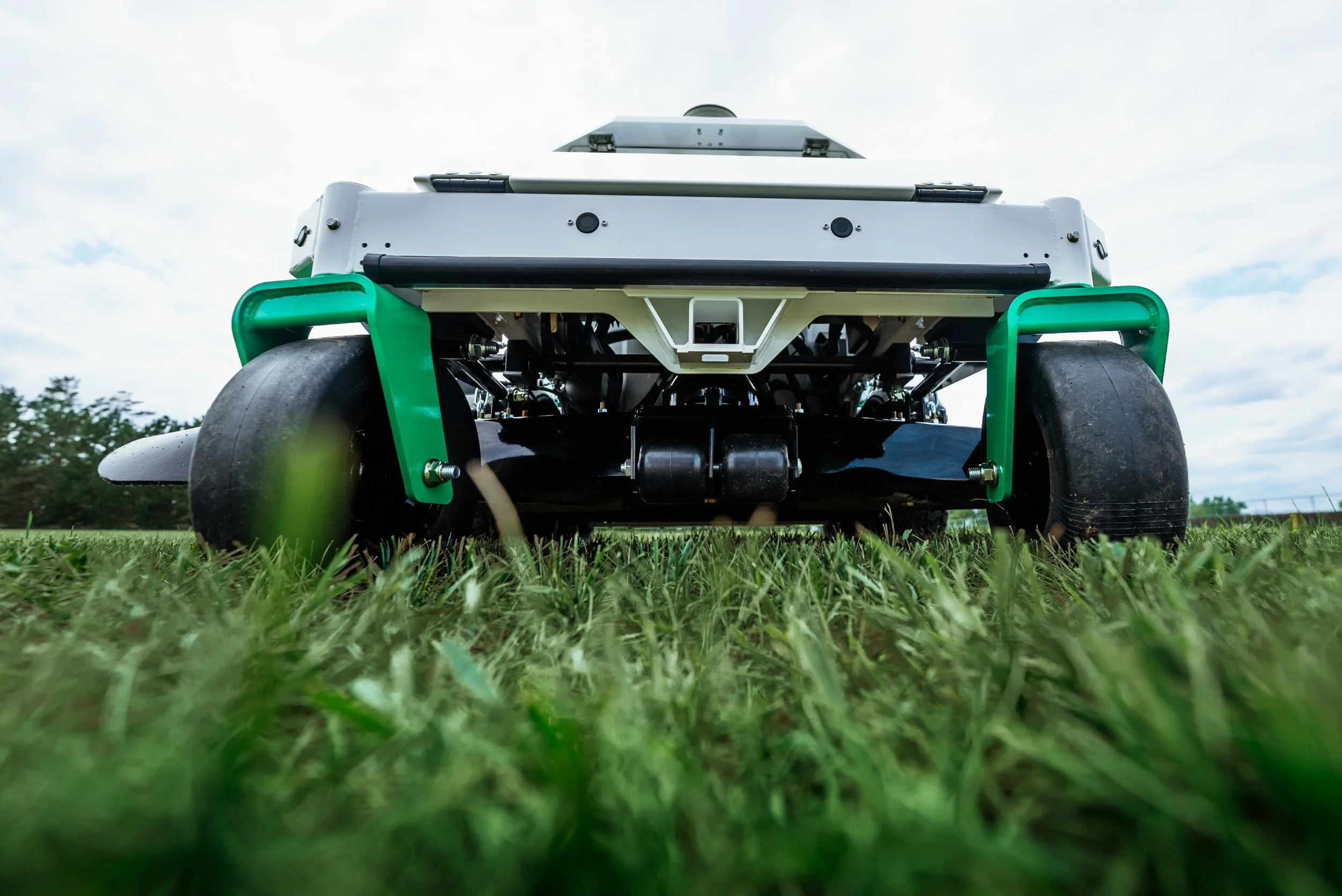 Close-up of an autonomous robot or drone with large wheels on a grassy field, taken from a low angle view with trees and cloudy sky in the background.