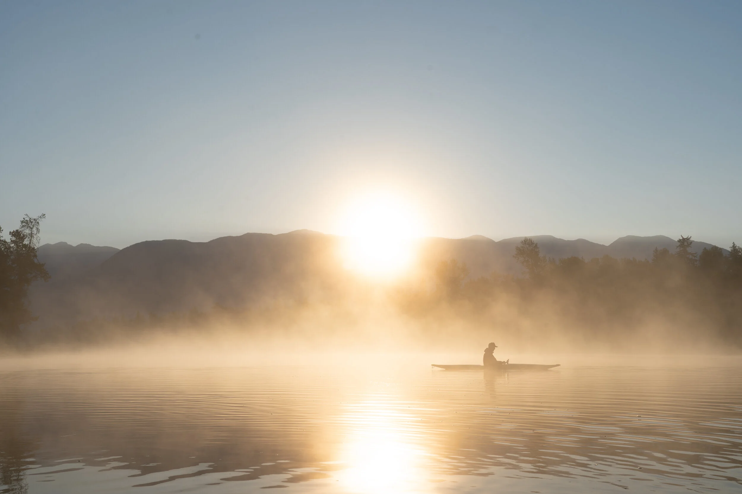 Person kayaking on a calm river at sunrise with mist and mountains in the background.
