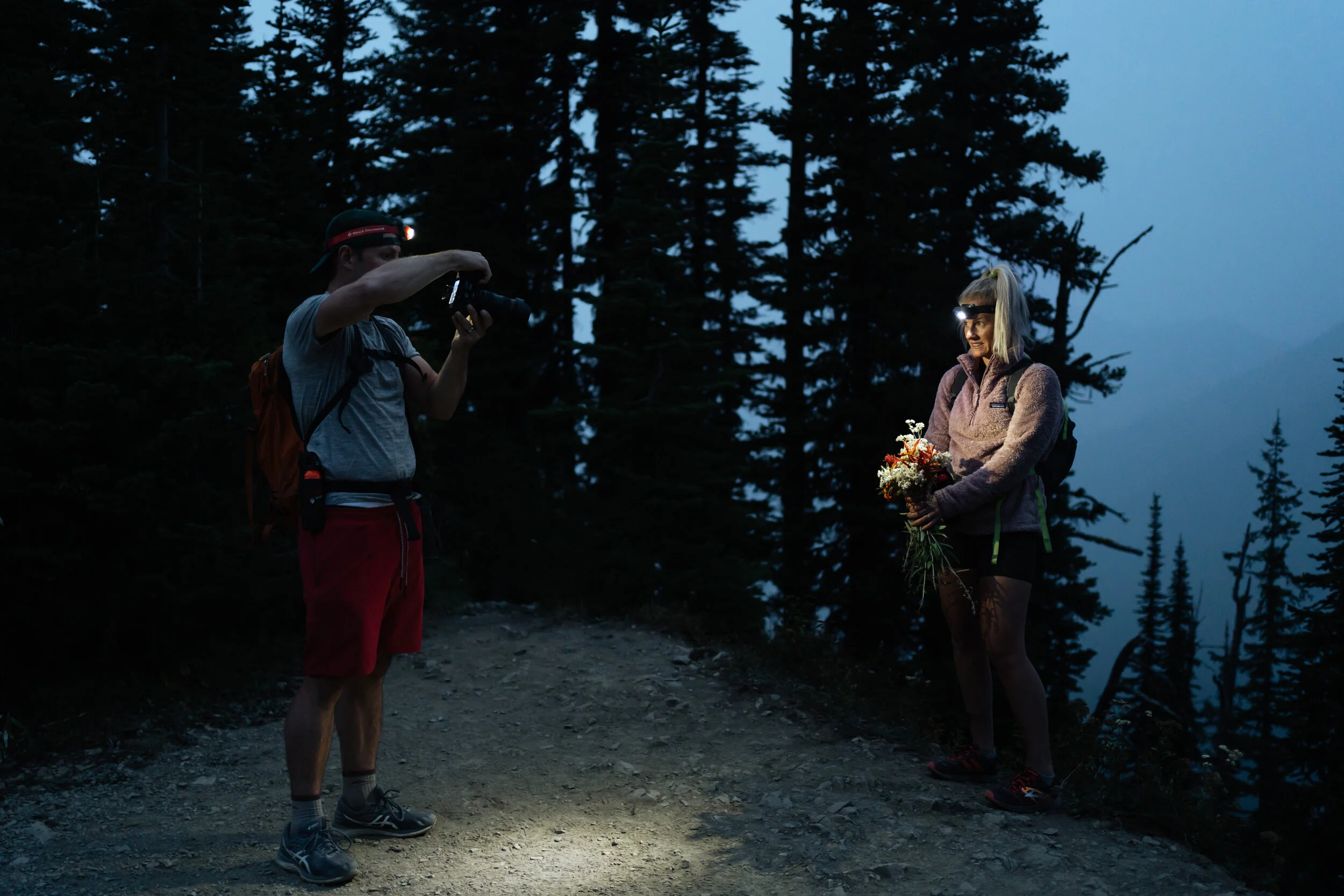 A woman standing on a mountain trail at dusk, holding a bouquet of flowers, being photographed by a man using a camera with a headlamp, surrounded by tall pine trees.