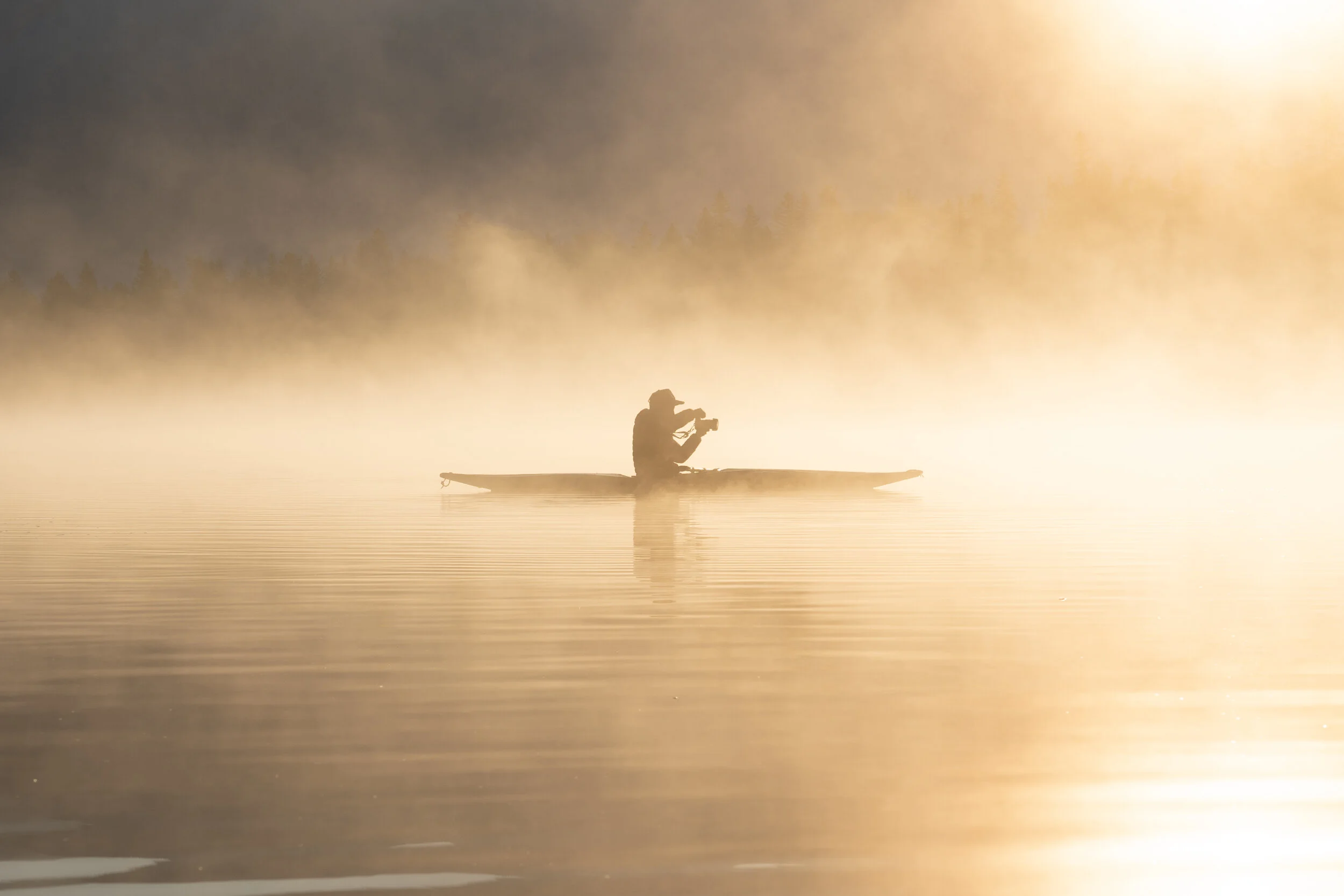 A person in a kayak on a calm body of water with mist and sunlight in the background.