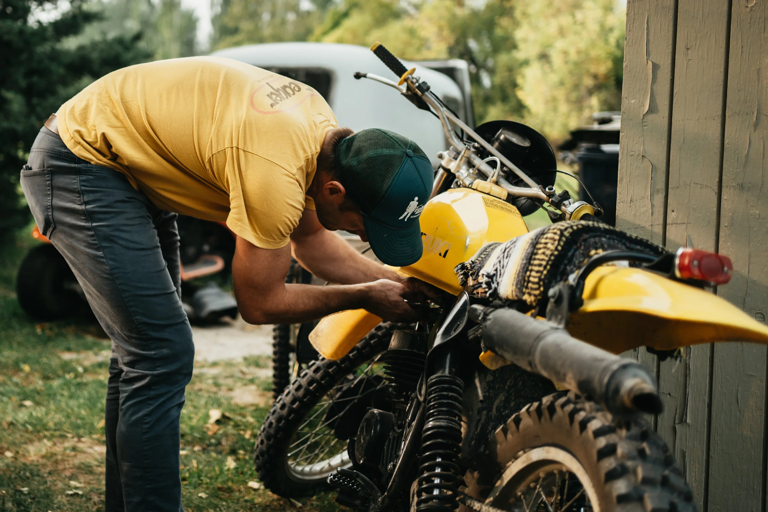 A man in a yellow shirt and green cap working on a yellow dirt bike outdoors near a wooden shed.
