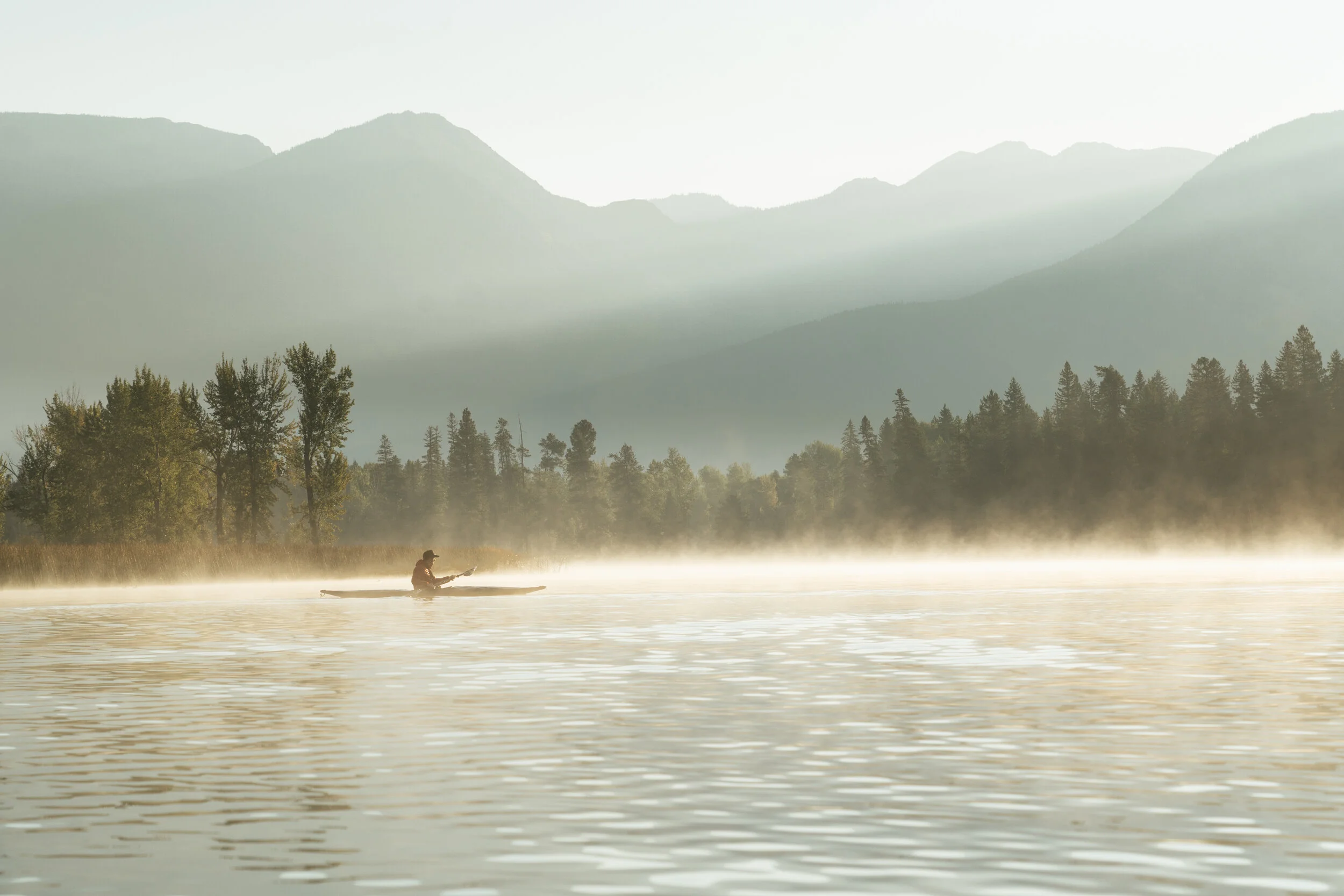 A person kayaking on a lake with a tree-lined shoreline and mountains in the background, early in the morning with mist rising from the water.