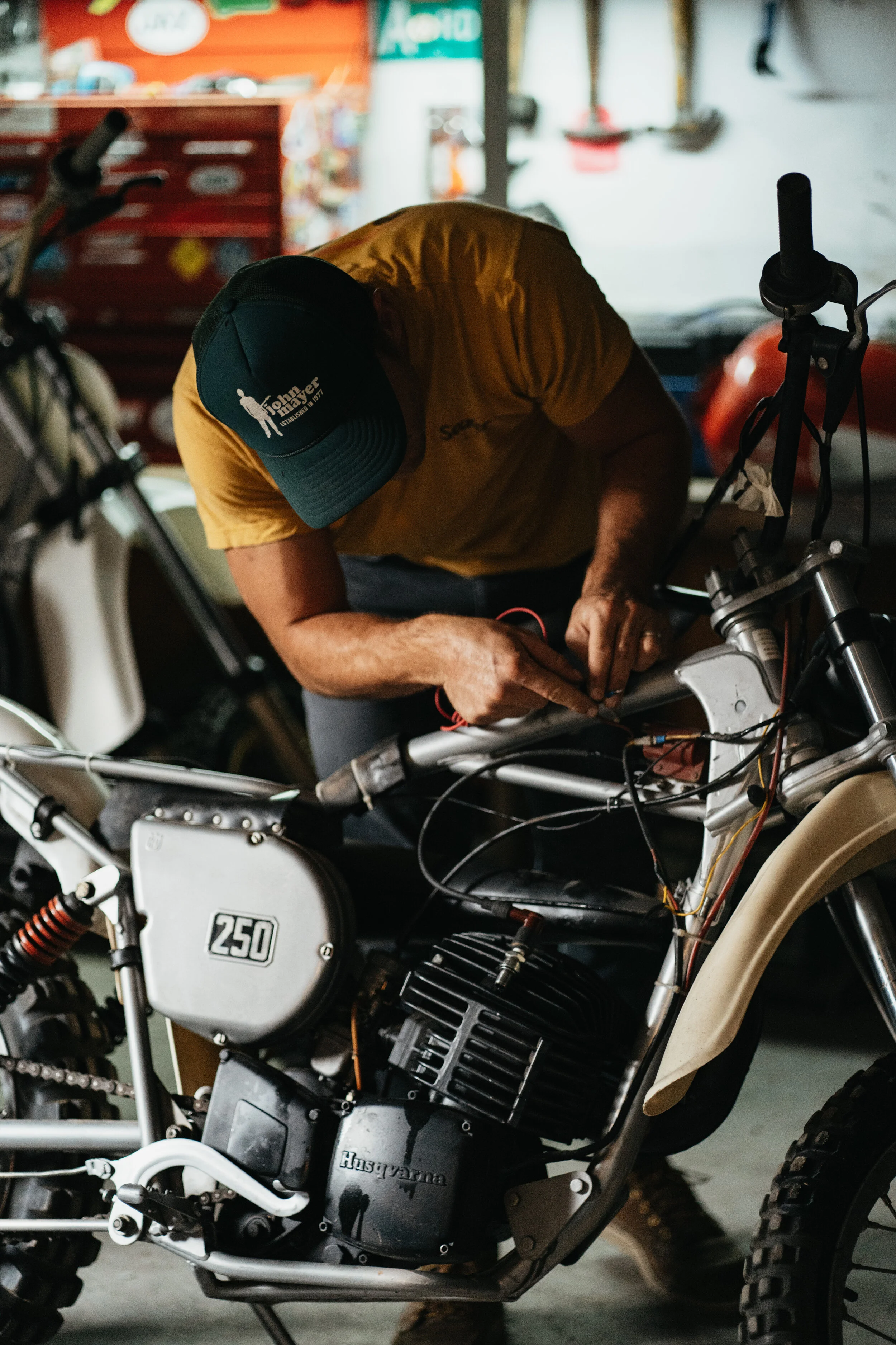 A man working on a vintage Husqvarna motorcycle in a garage, wearing a green cap and a yellow T-shirt, with tools and other motorcycles in the background.