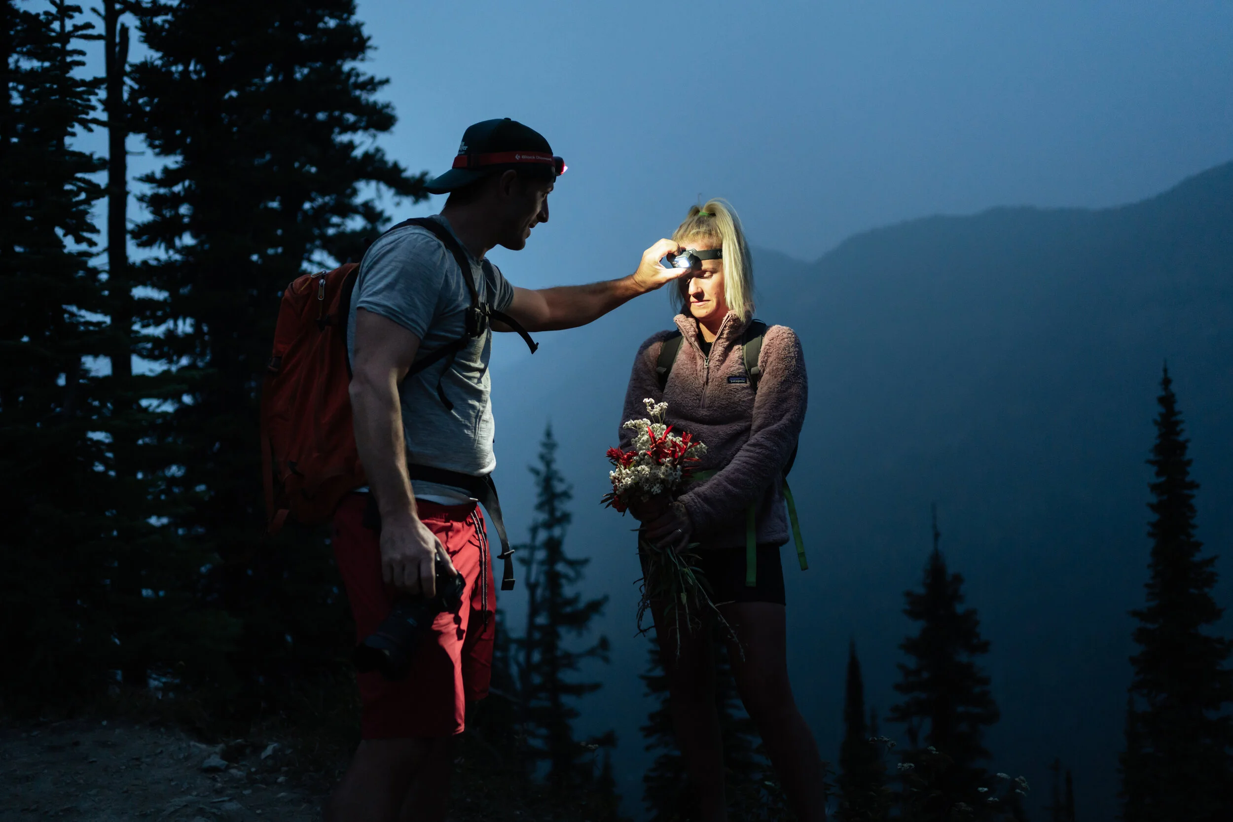 A man giving a woman flowers on a mountain trail during dusk, with trees and mountains in the background.