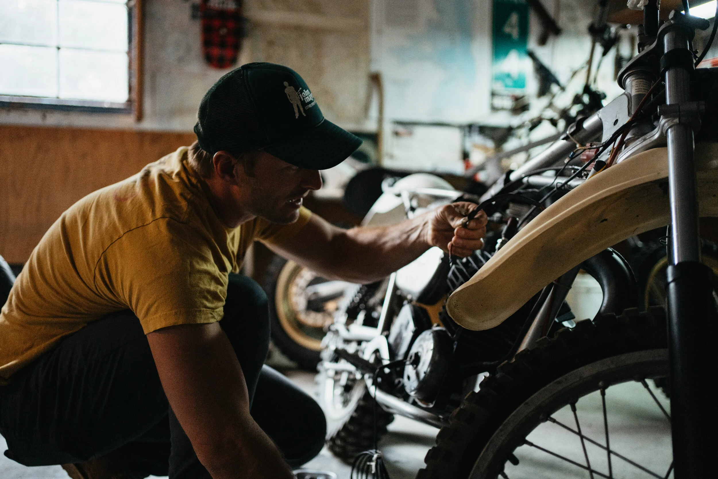 A man wearing a yellow T-shirt and a black cap working on a motorcycle in a garage.