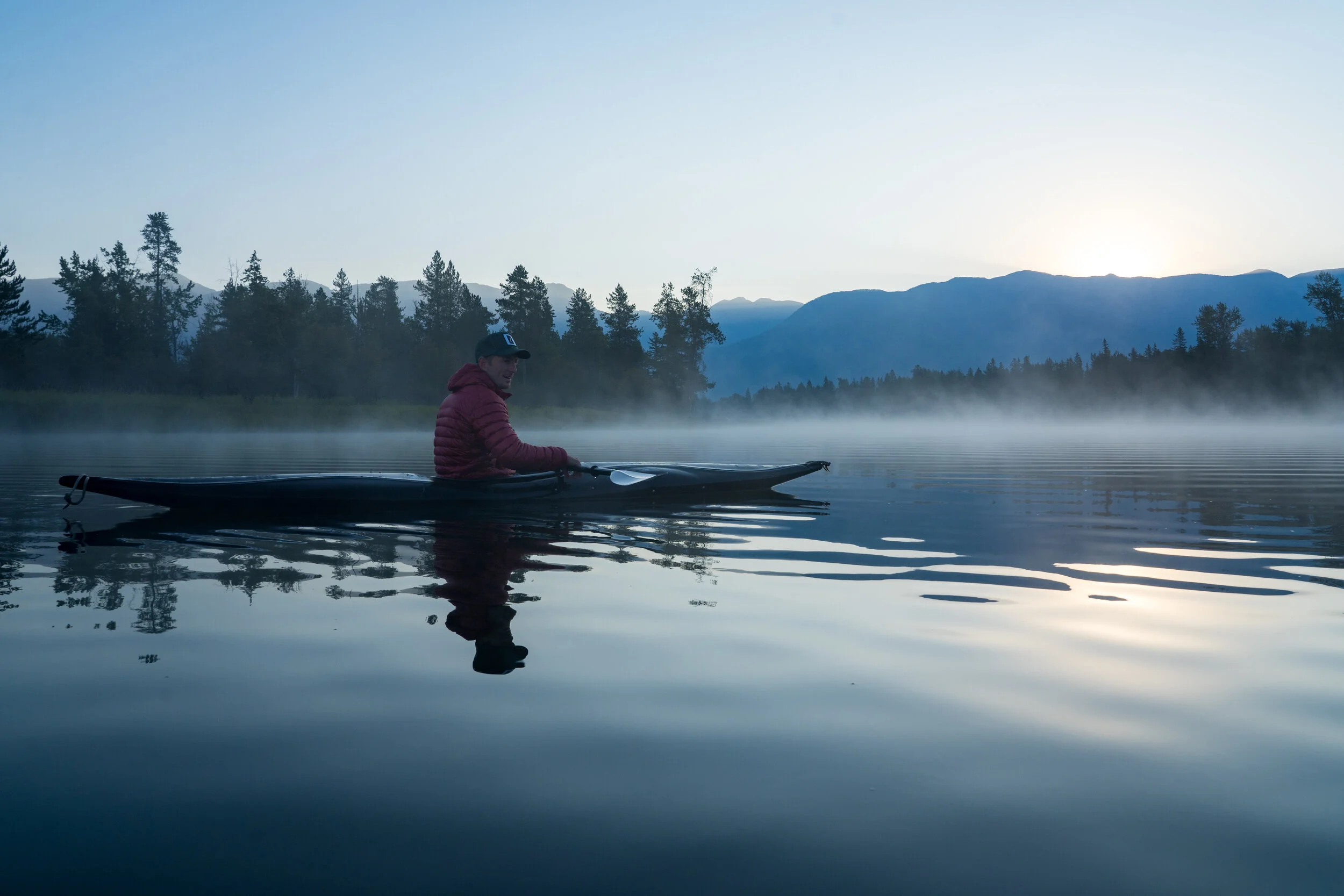 A person in a red jacket and black cap sitting in a kayak on a calm lake at sunrise or sunset, with mist rising from the water and mountains in the background.