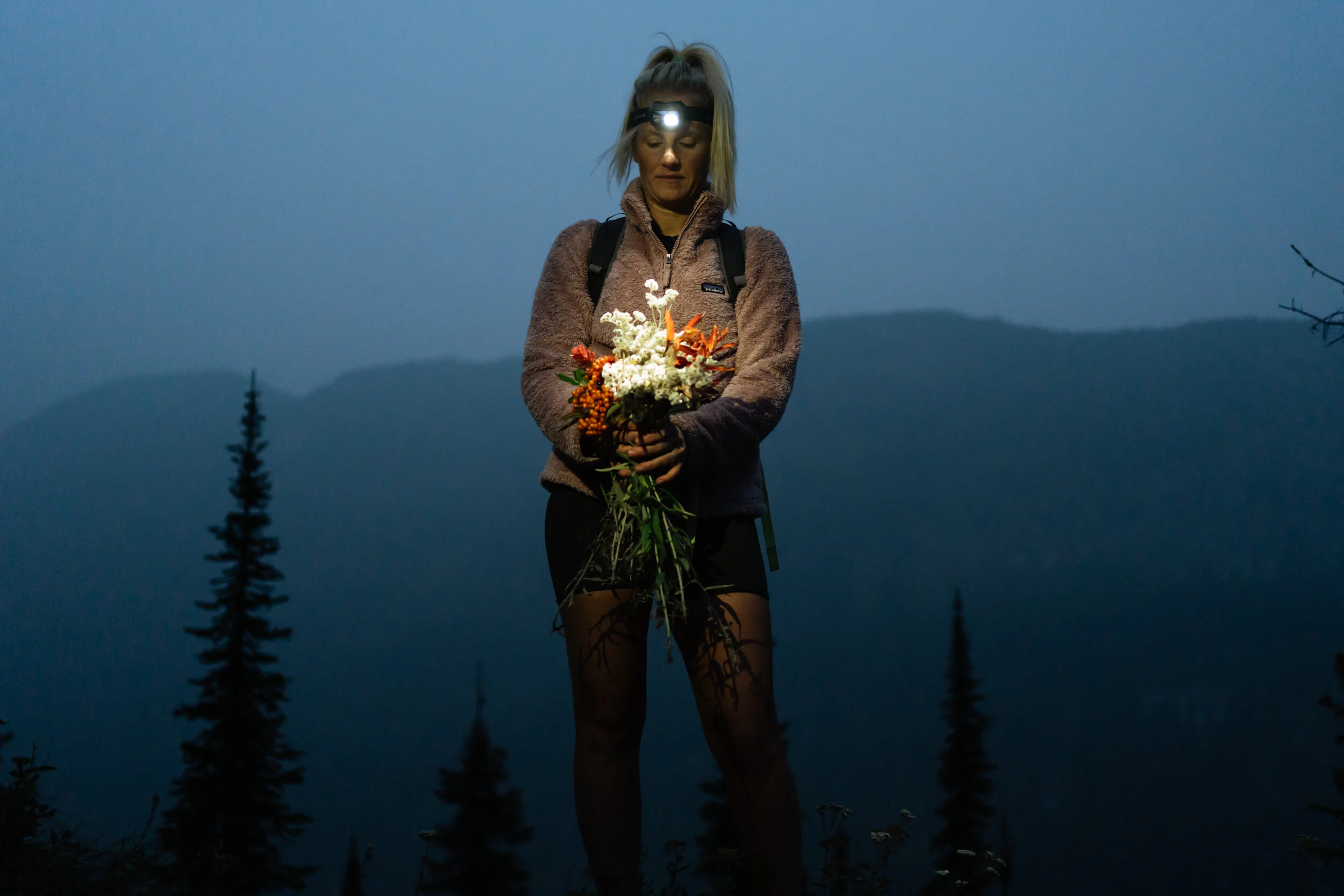 A woman wearing a headlamp and outdoor clothing holding a bouquet of flowers at dusk with a mountain and pine trees in the background.