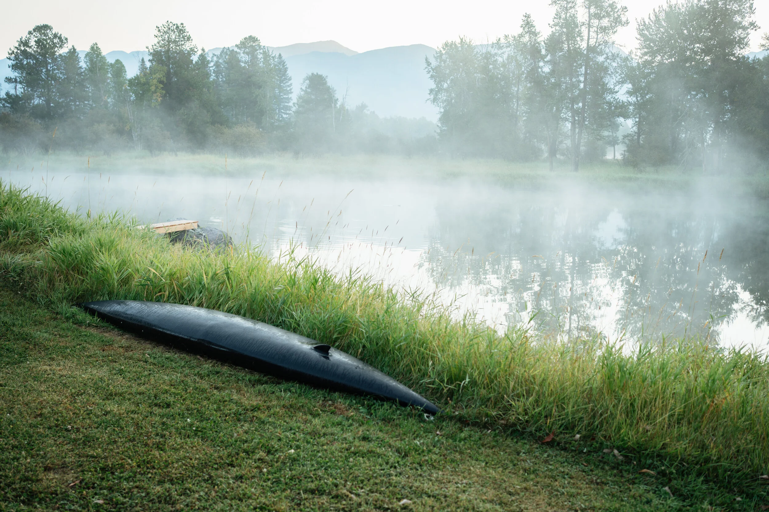 A black canoe resting on the grassy shore of a foggy river with trees and mountains in the background.