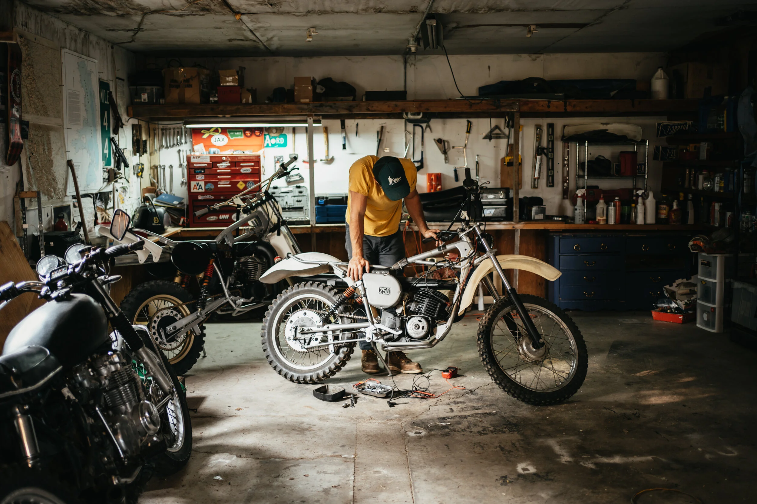 A person working on a vintage off-road motorcycle in a cluttered garage, with tools hanging on the wall and additional motorcycles parked around.