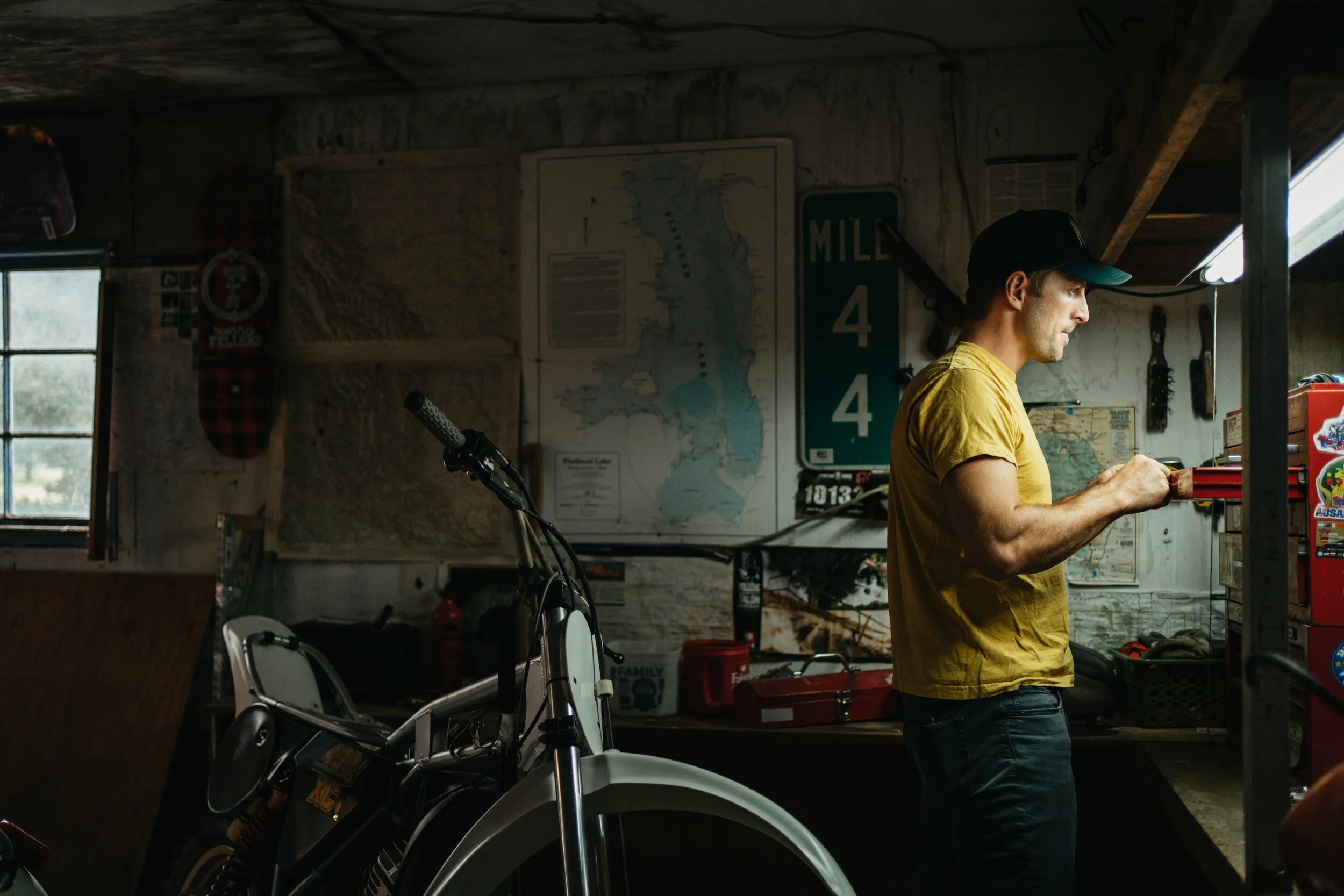 A man in a yellow shirt and black cap working on a red tool in a garage with a motorcycle, wall maps, and sports memorabilia.