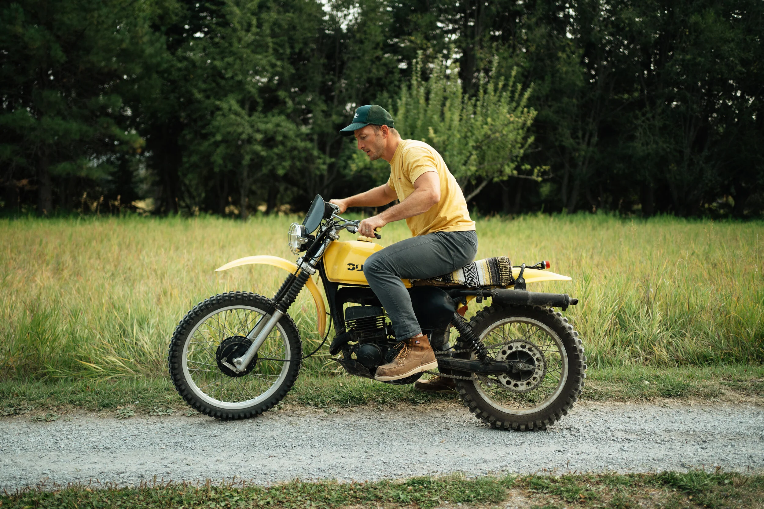 A man riding a vintage yellow motorcycle on a gravel path through a grassy field with trees in the background.