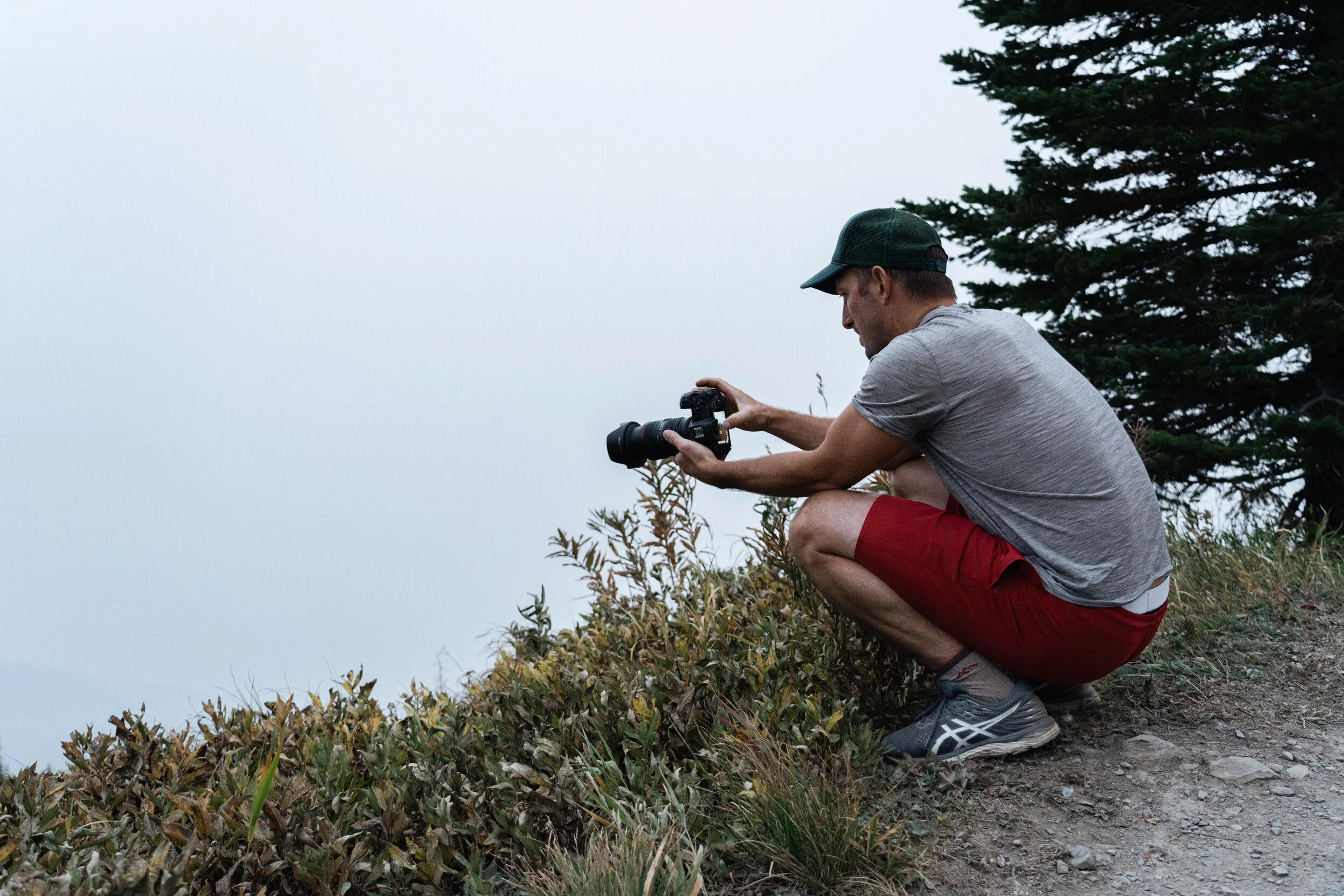 A man crouching on a mountain trail, holding a camera and preparing to take a photograph of the fog-covered landscape with trees in the background.