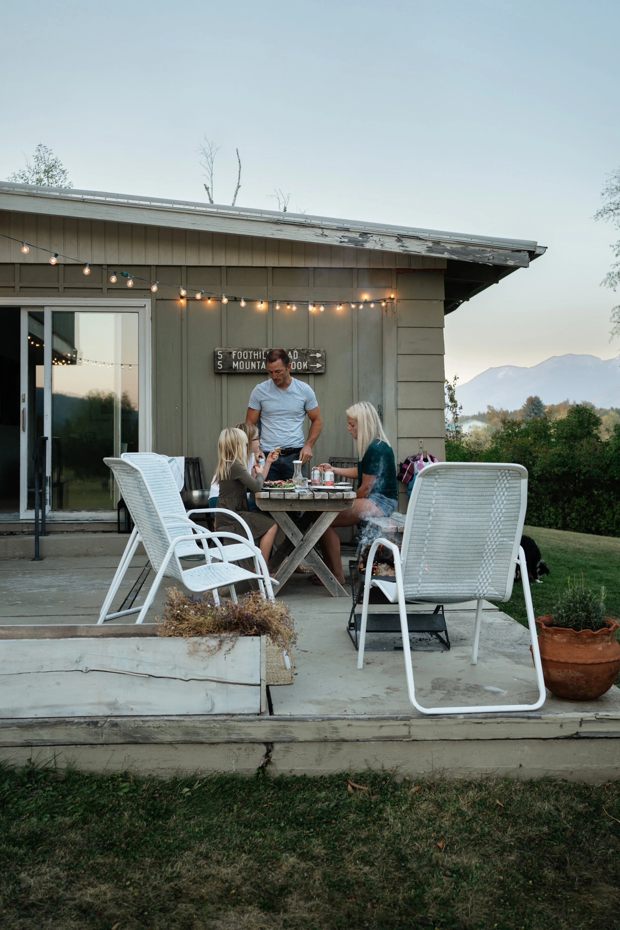 A family of four, with three women and one man, sitting at a wooden outdoor dining table on a porch, grilling food with a small charcoal grill. The home has string lights hanging from the roof and a mountain view in the background.