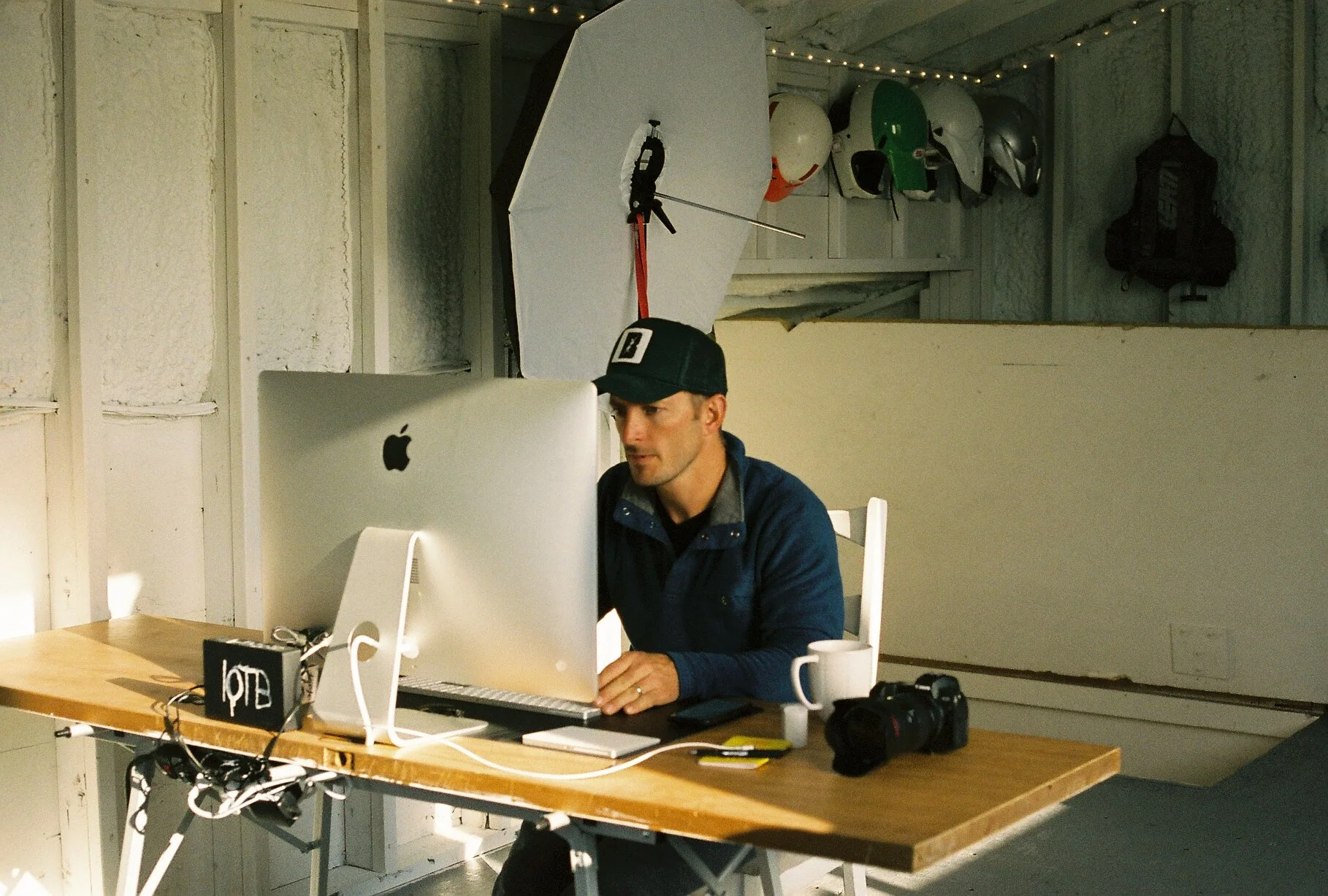 A man working at a desk with a large iMac computer, wearing a black cap and a blue jacket. Behind him, there are several helmets and a couple of bags mounted on the wall. On the desk, there are a camera, a coffee mug, and some electronic items, with 