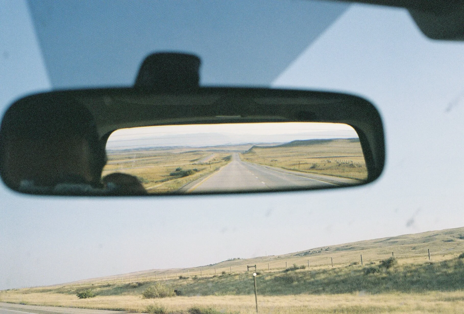 A view of a straight highway through a flat, open landscape, as seen through a car's rearview mirror.
