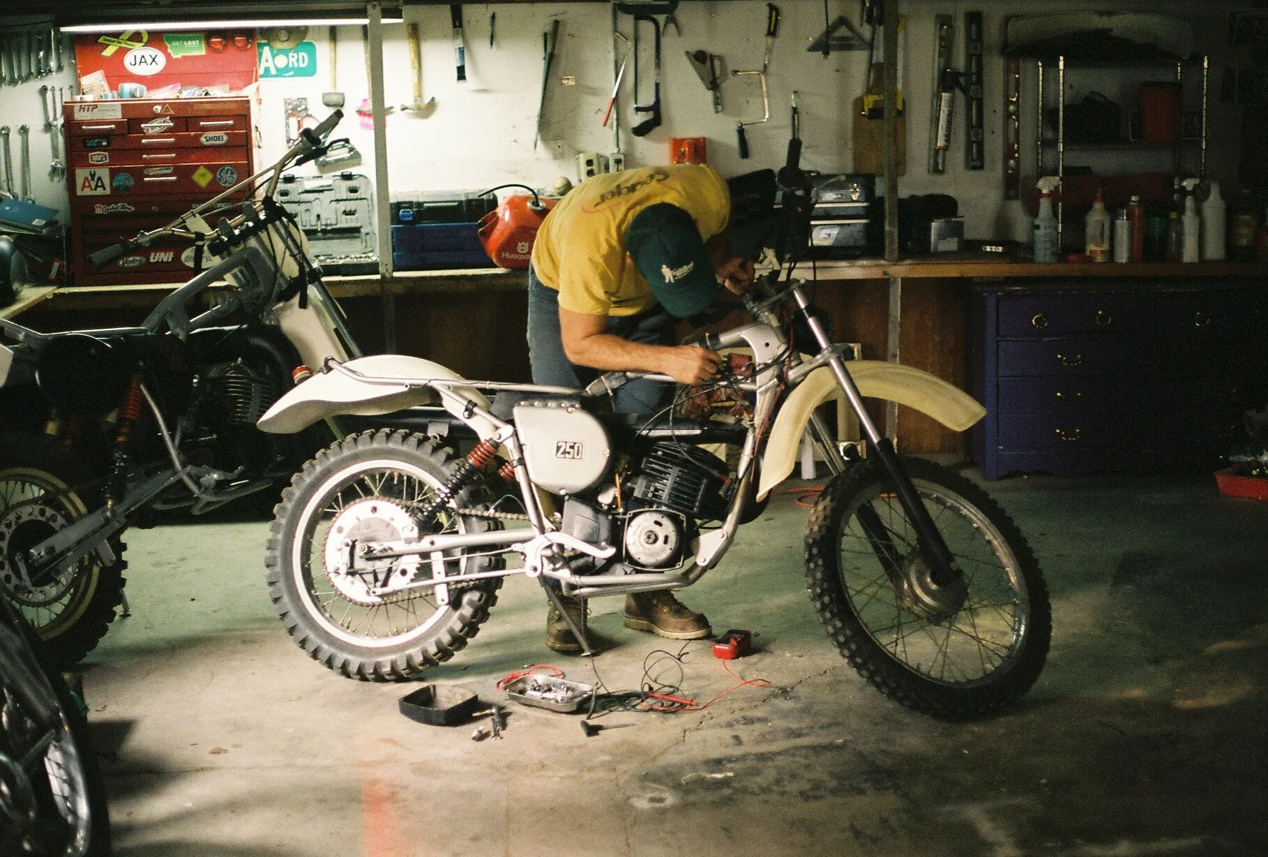 A person working on a vintage dirt bike in a garage, with tools and equipment hanging on the wall and a toolbox in the background.
