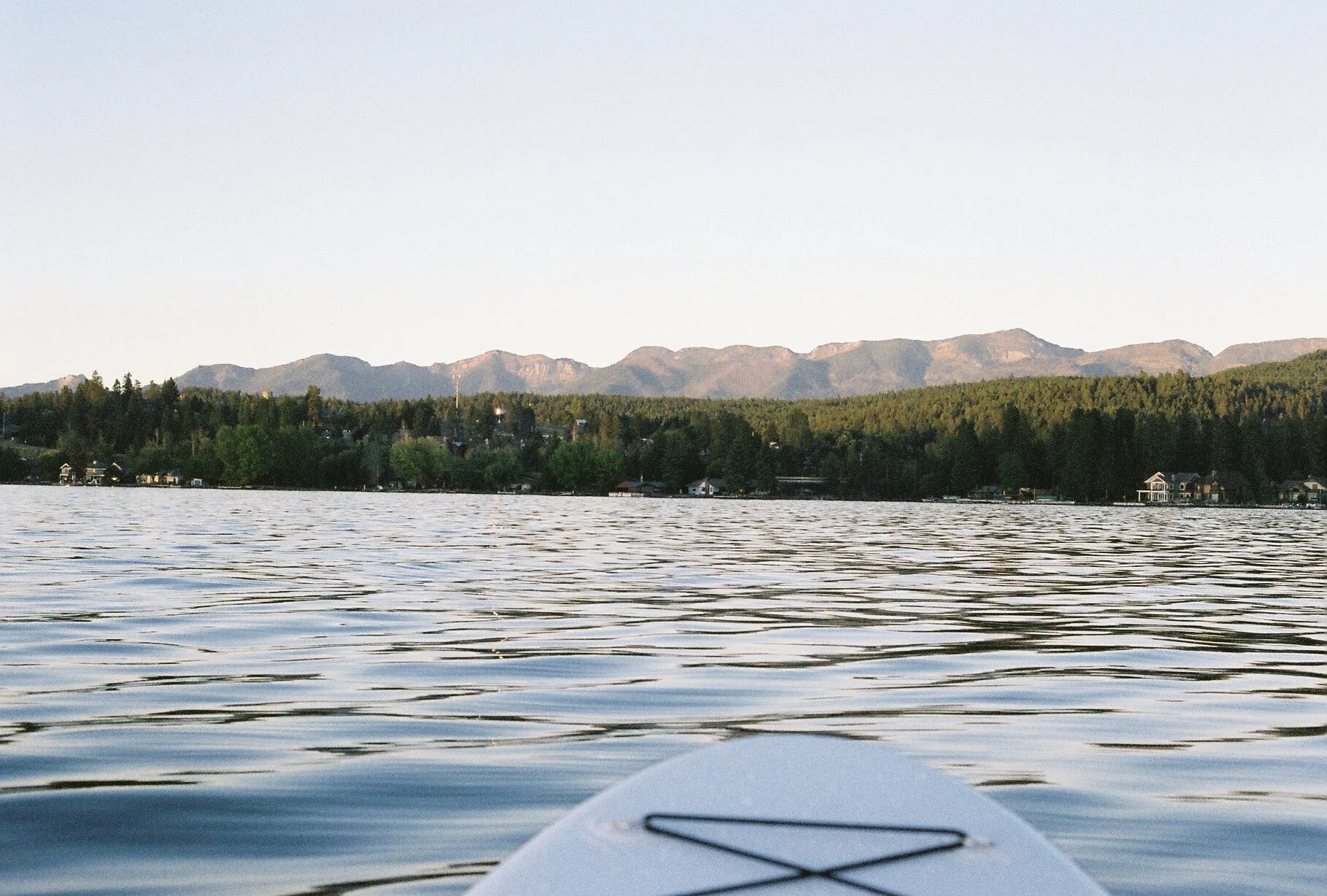 View from a kayak on a lake with houses along the shoreline, surrounded by trees and distant mountains under a clear sky.