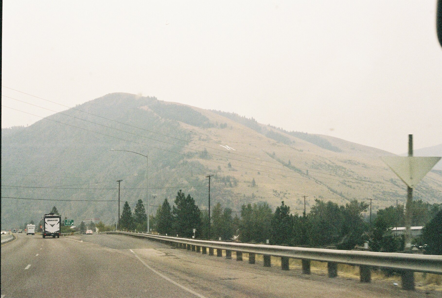 A highway with vehicles driving, surrounded by trees, and a large mountain in the background in a hazy sky.