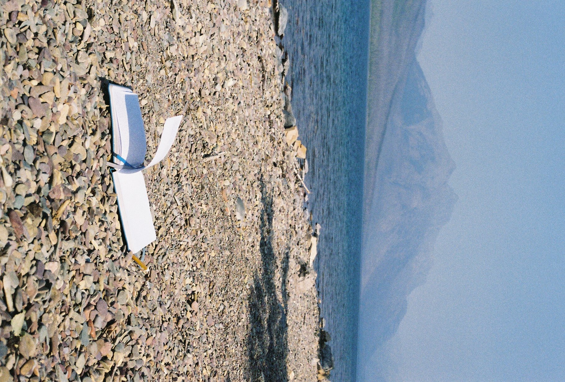 Notebook, pen, and a filter placed on a pebbled beach with water and mountains in the background.