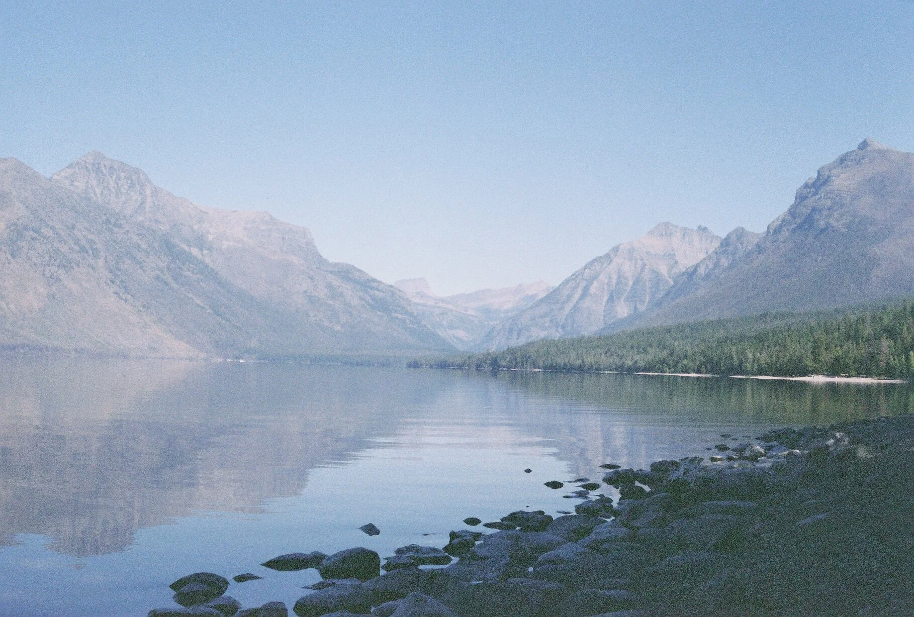 A calm lake reflecting mountains with snow-capped peaks in the distance, a rocky shoreline in the foreground, and a tree line along the far edge of the water.