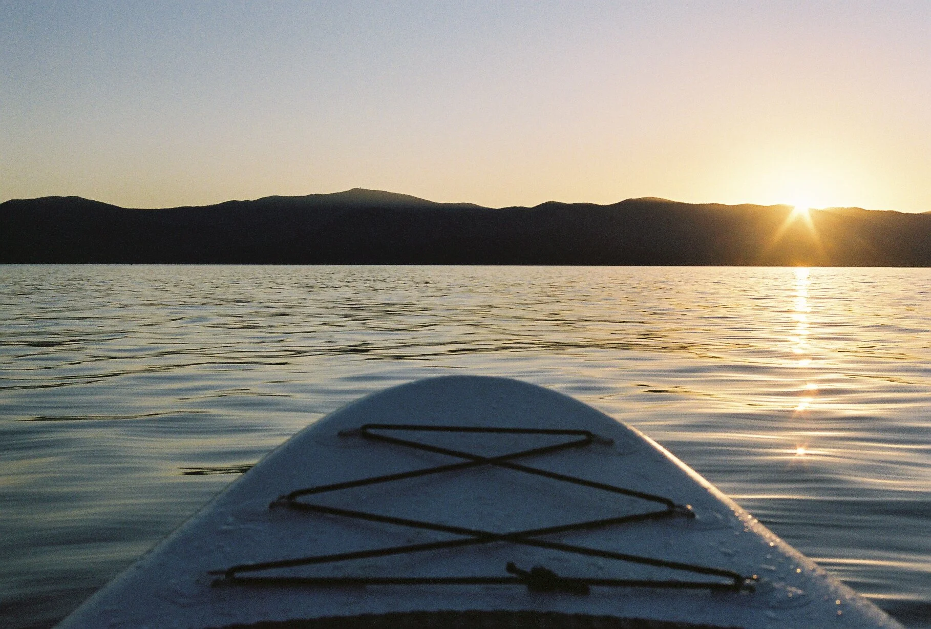 View from a kayak or paddleboard on a calm body of water during sunset, with distant hills on the horizon and the sun partly visible near the right side.