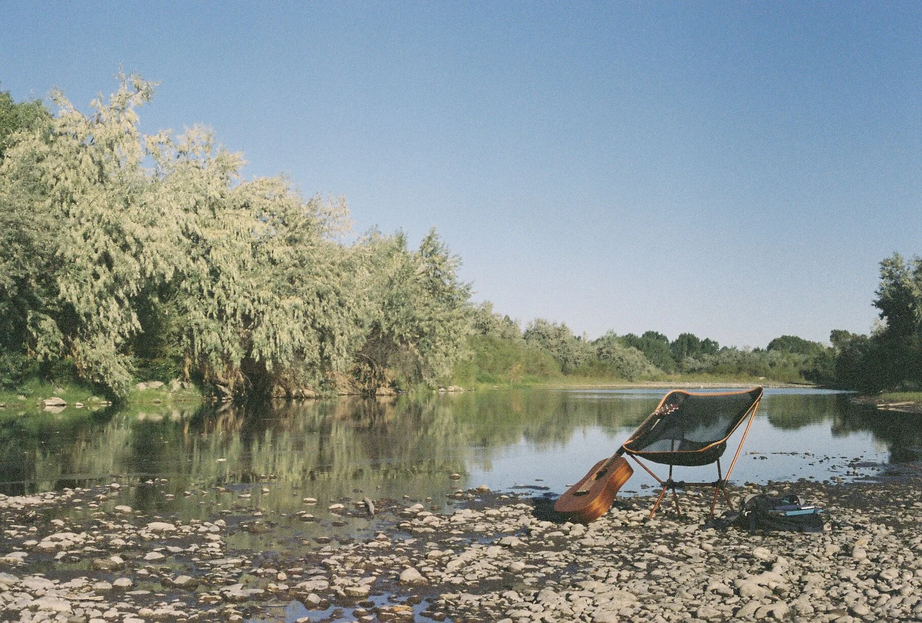 A river scene with a chair and a guitar on rocky riverbank, trees and blue sky reflected in the water.