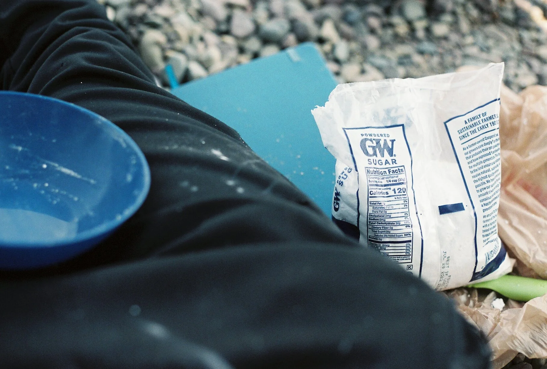 Close-up of a bag of powdered sugar resting on a person's lap, with a blue container and a blue folder or notebook nearby. The person is wearing black pants, and the background features a rocky ground surface.