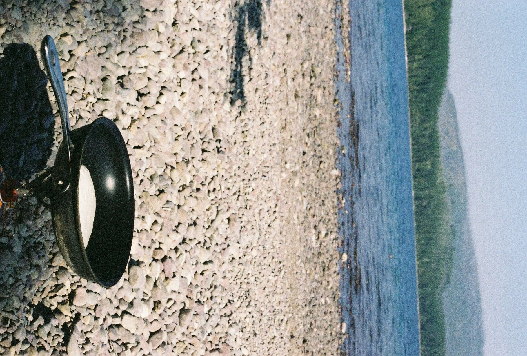 A black frying pan with a white substance, possibly batter, on a rocky shoreline with water and distant hills in the background.