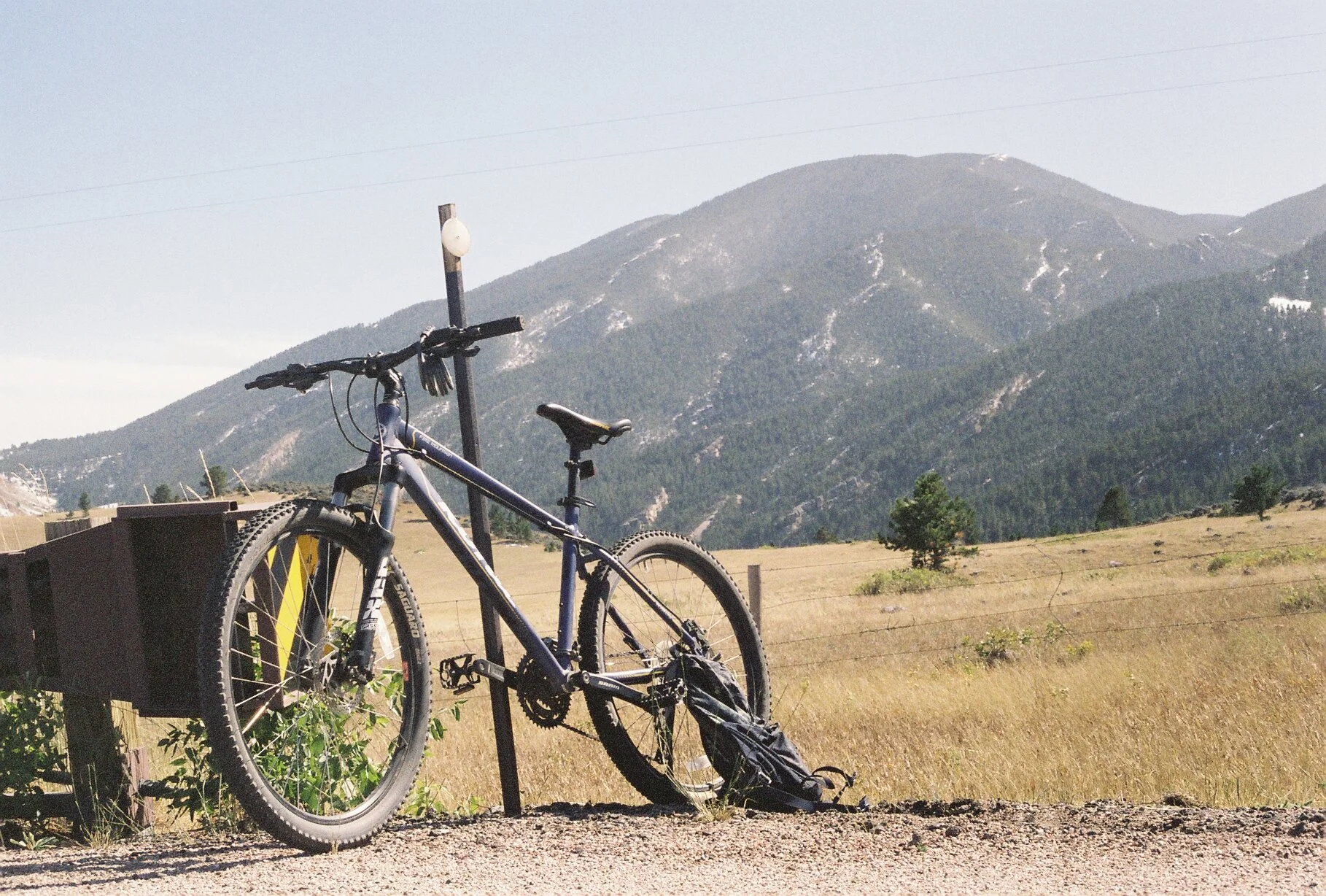A mountain bike leaning against a post with a backpack on the ground in a rural landscape with mountains in the background.