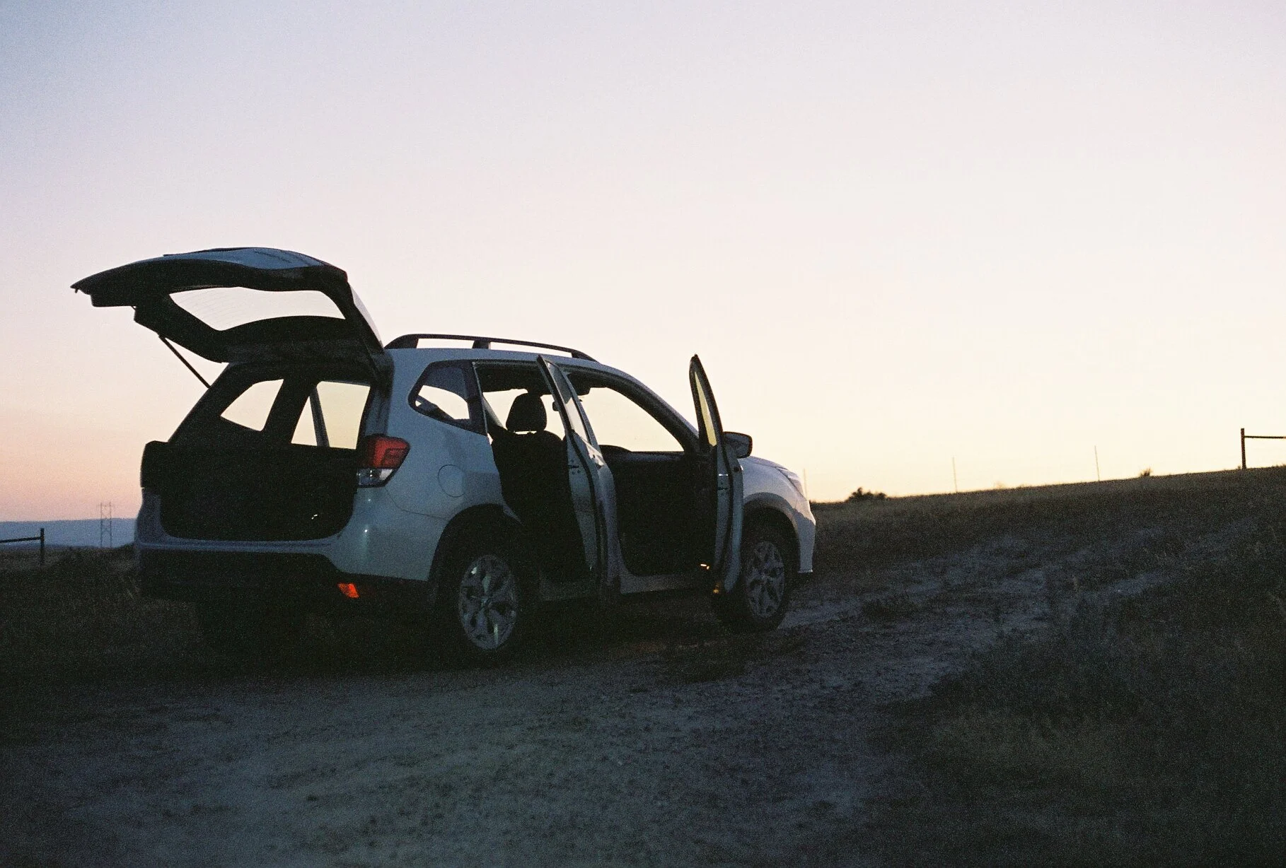 White SUV with open doors and trunk parked on a dirt road during sunset or sunrise.