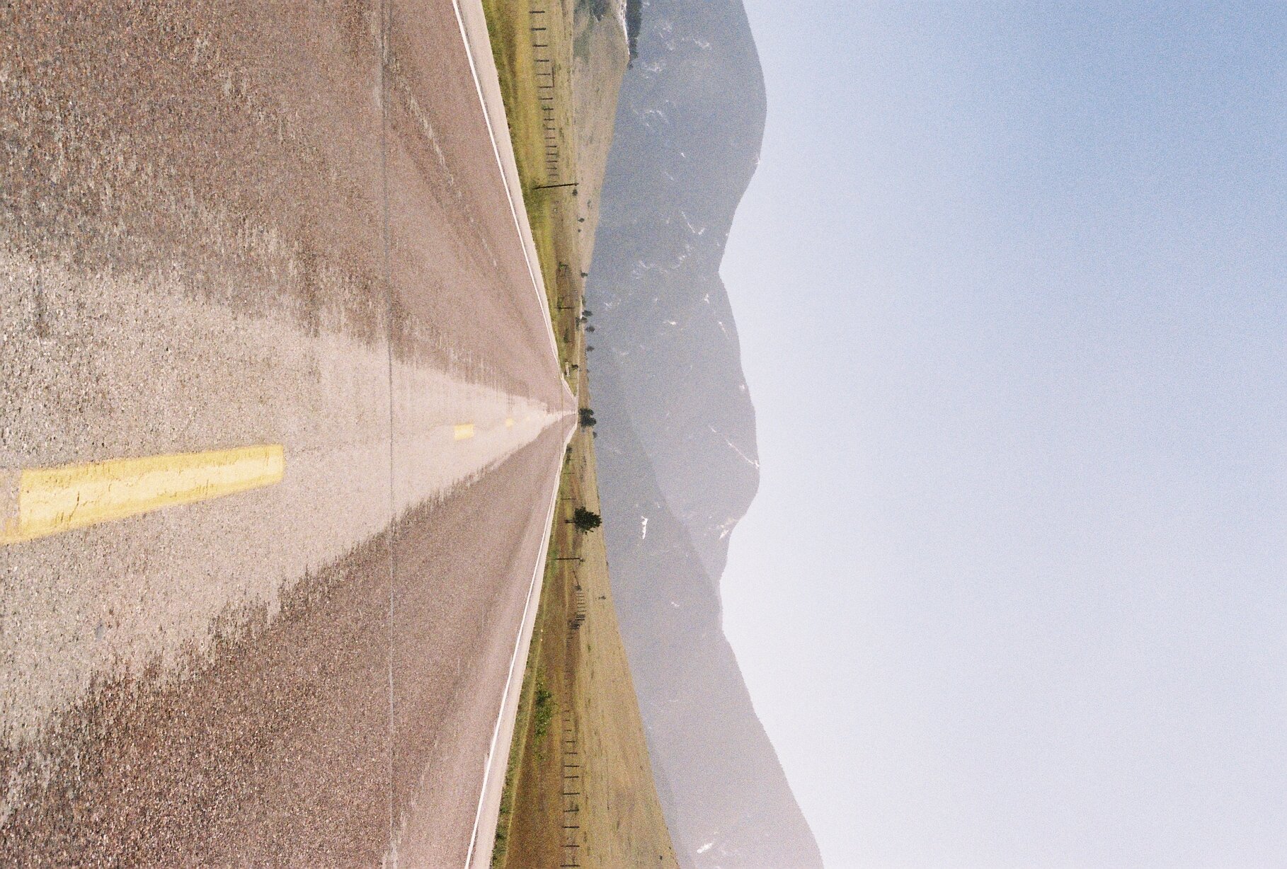 Empty two-lane road stretching into the distance with mountains in the background, under a clear or slightly hazy sky.