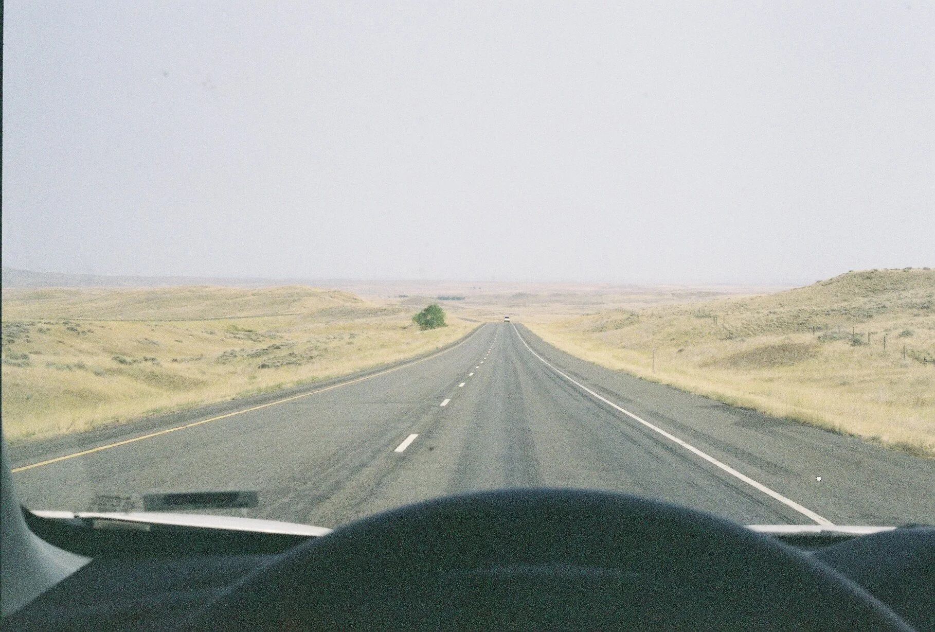 View from inside a vehicle showing a two-lane highway stretching through a flat, open landscape with dry grass and a few small hills under a pale, overcast sky.
