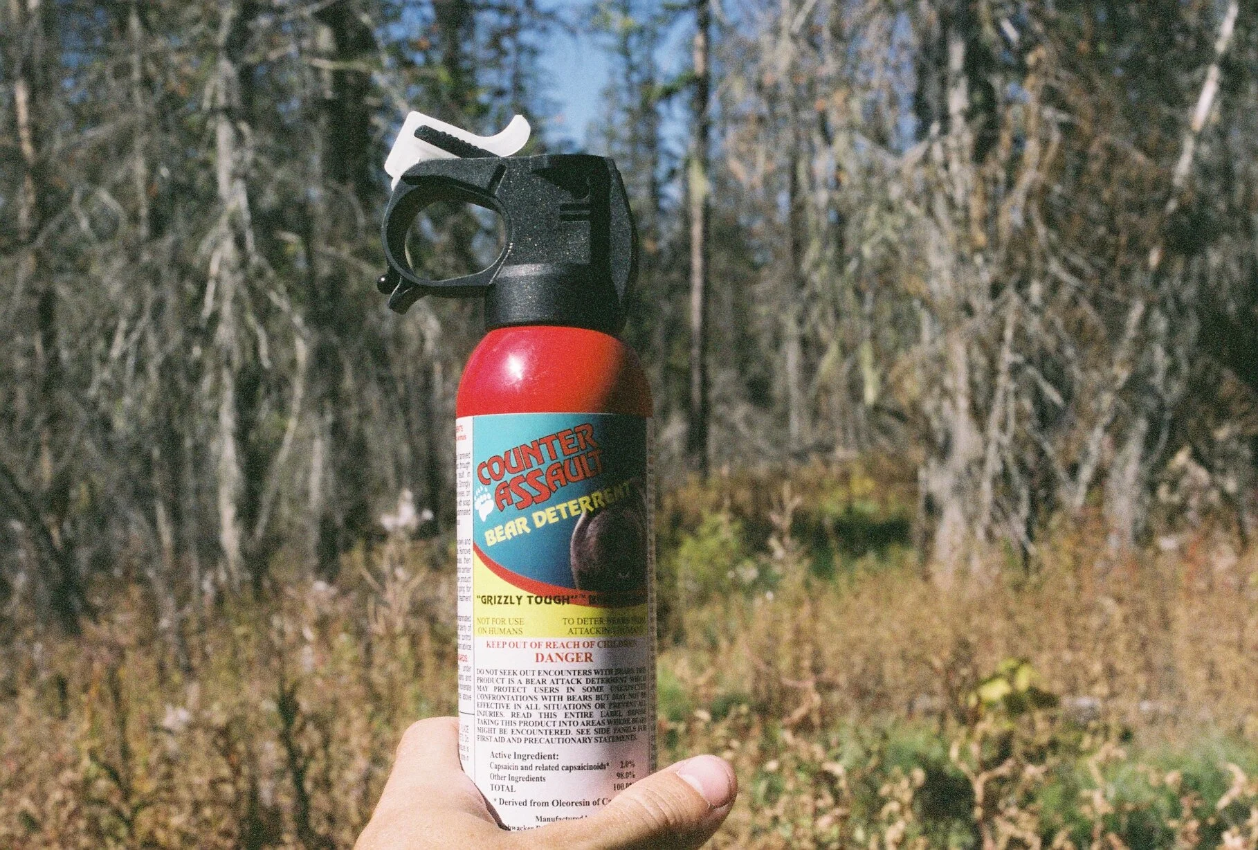 A person holding a bear deterrent spray with a spray nozzle, outdoors in a forested area with trees in the background.