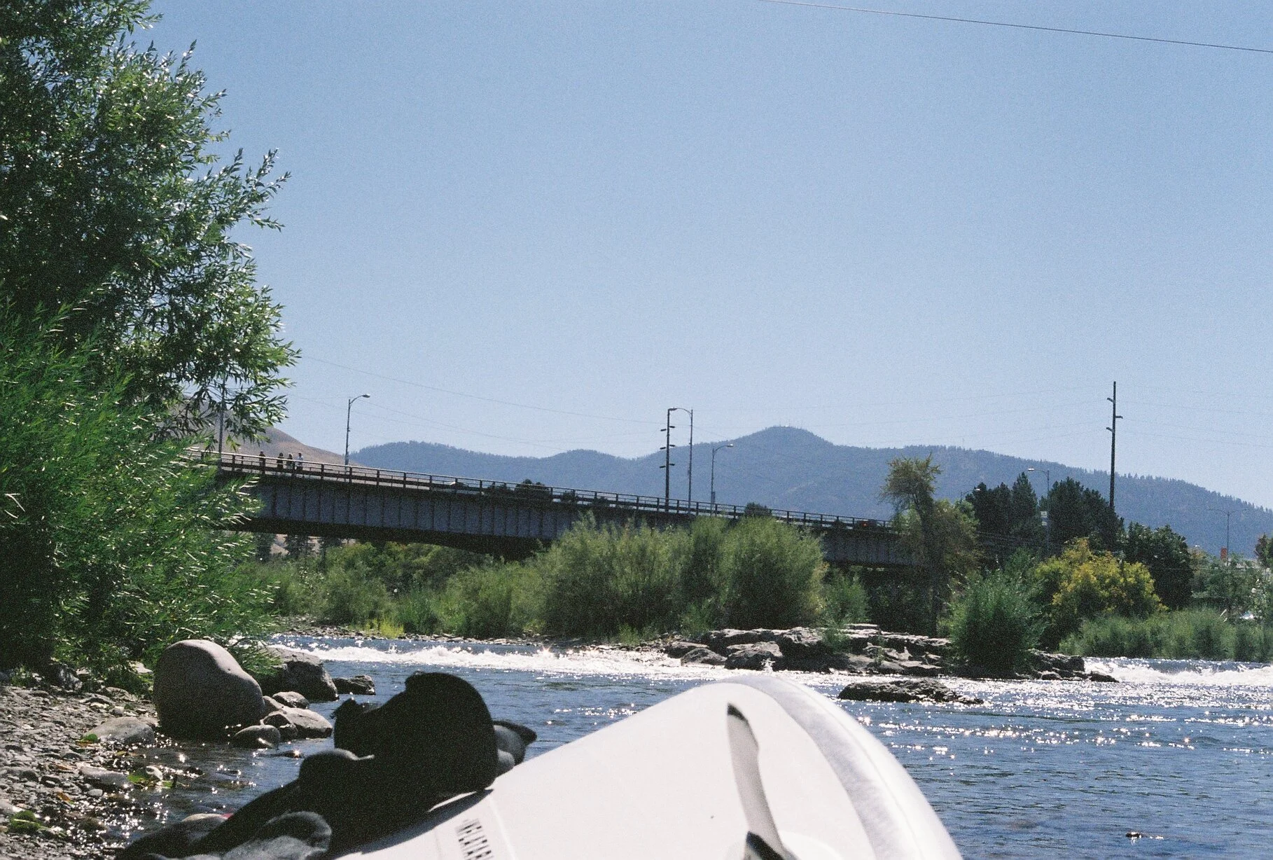 View of a river with rocks and trees on the bank, a bridge in the background, and mountains under a clear sky.