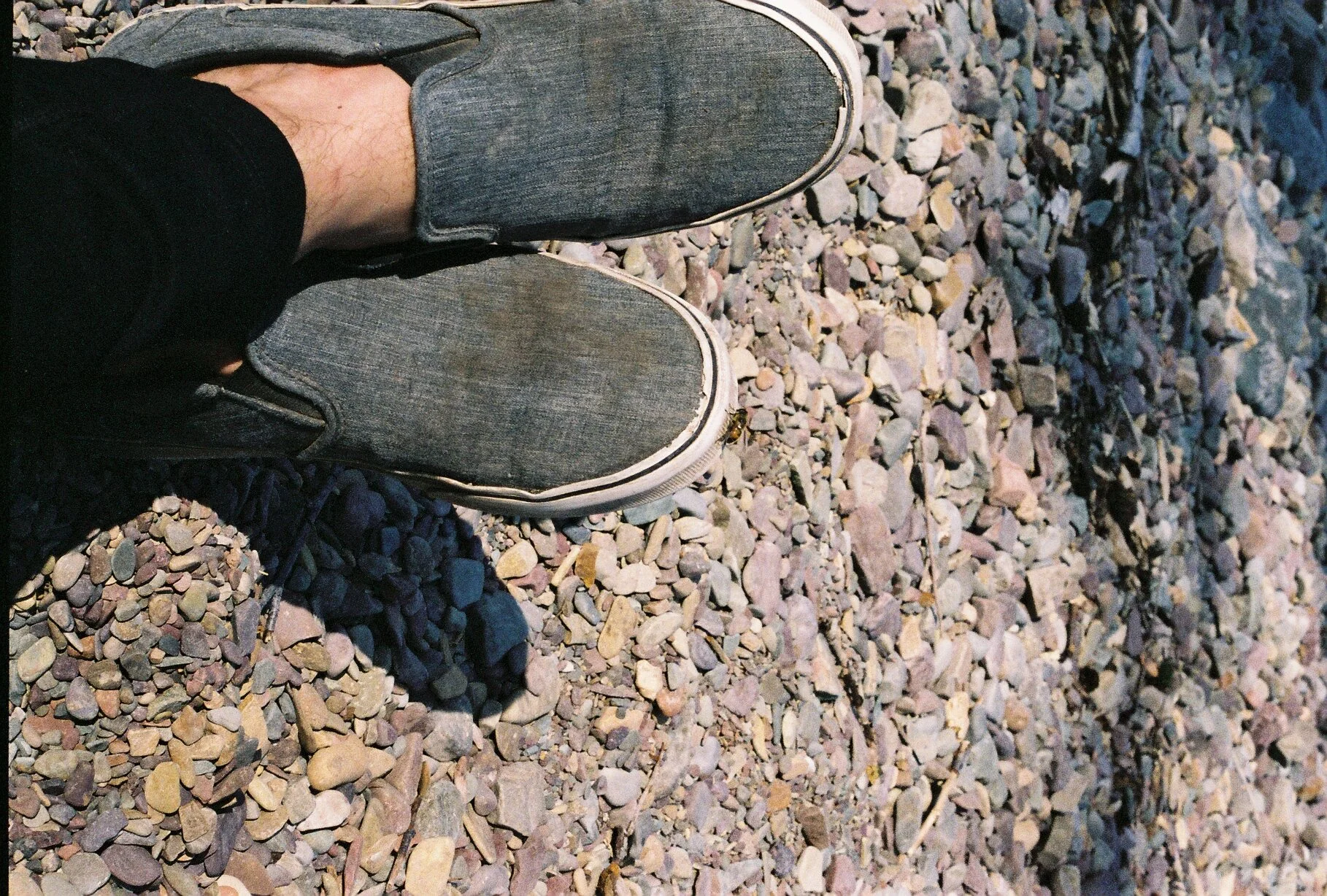 A person's legs wearing black pants and gray slip-on shoes, standing on a rocky surface with small mixed-colored stones.