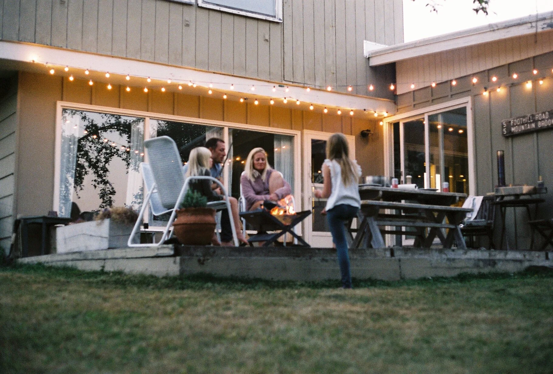 A family of four sitting around a firepit on a backyard patio, while a young girl stands nearby, with a house decorated with string lights in the background.