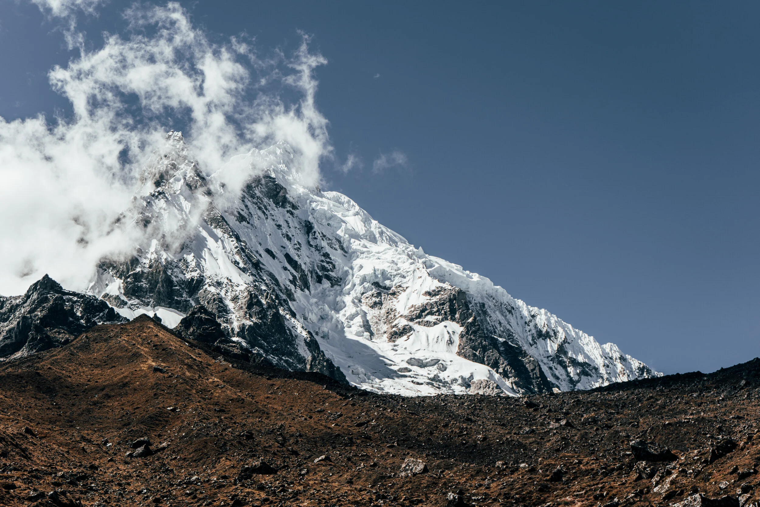 Snow-capped mountain peak with a brown rocky foreground and a blue sky.