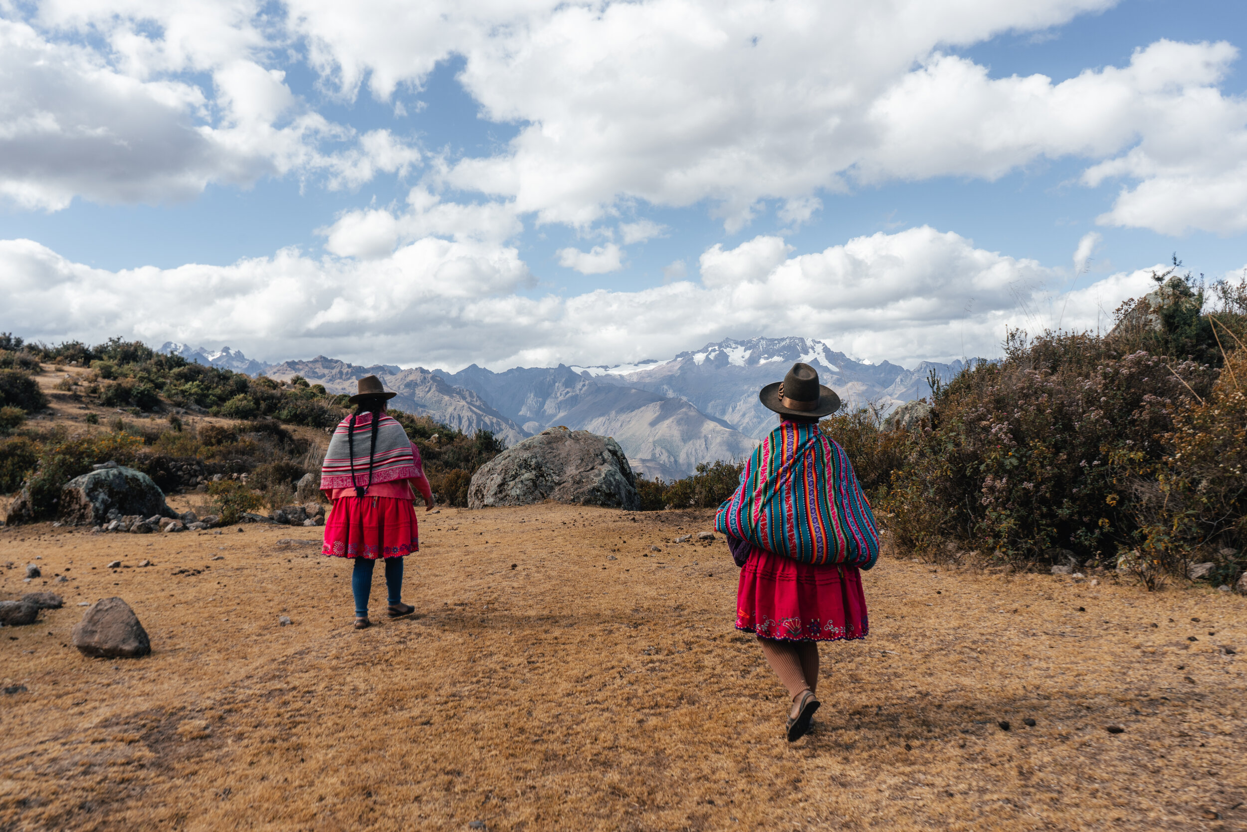 Two women dressed in colorful traditional Andean clothing walk through a mountainous landscape with clouds and snow-capped peaks in the background.