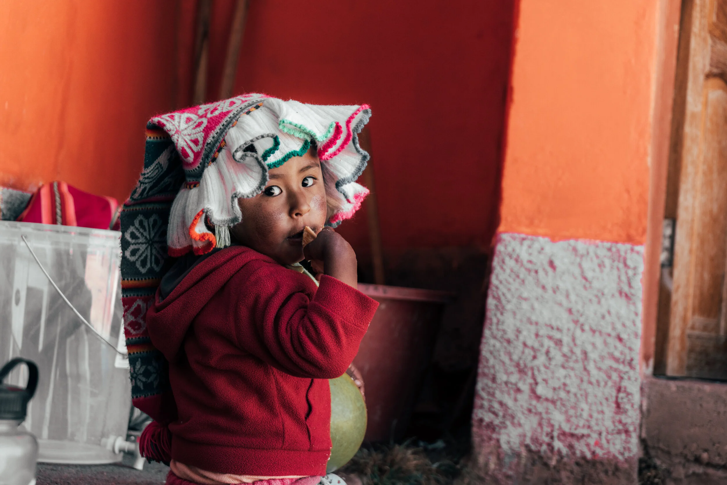 A young child with dark skin, wearing a colorful woolen hat and a red jacket, stands indoors near an orange wall, holding and eating a snack while looking towards the camera.