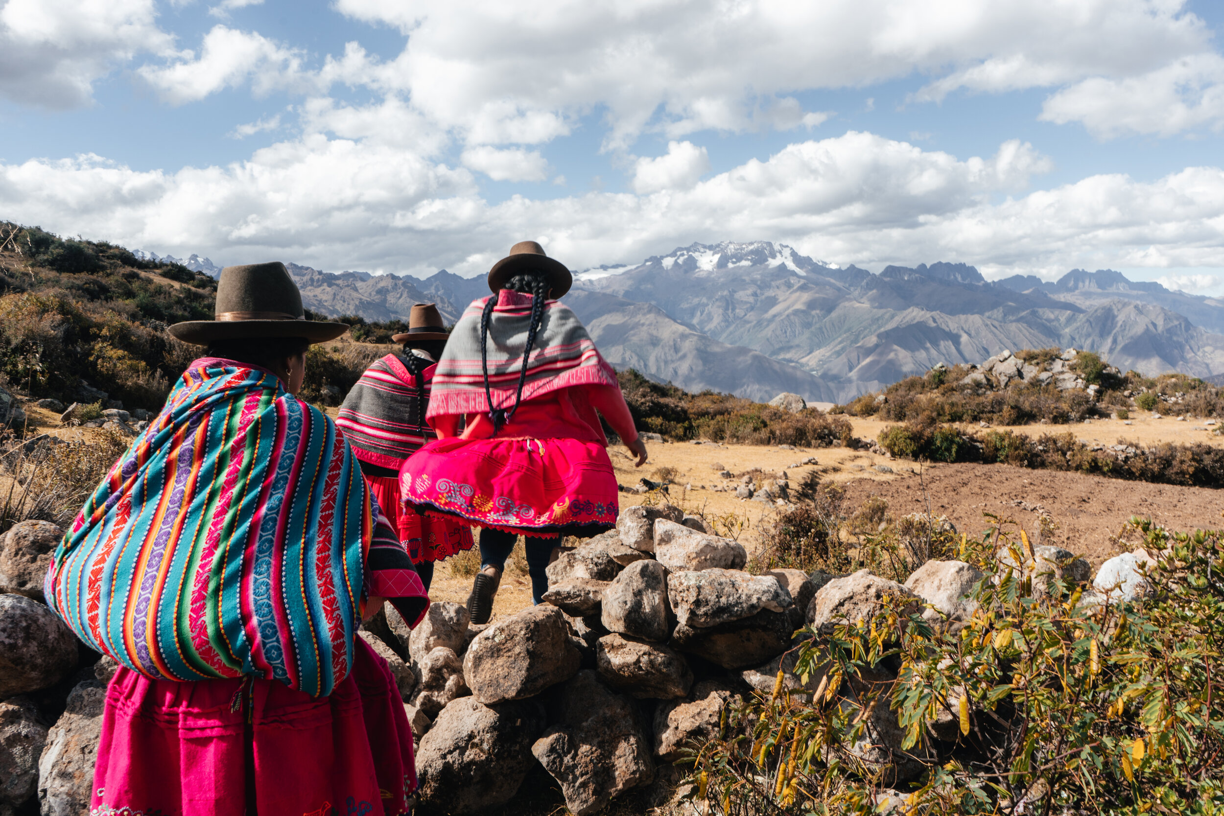 Three women dressed in traditional Andean clothing with colorful striped shawls and skirts, walking across a rocky landscape with mountains in the background and a partly cloudy sky.